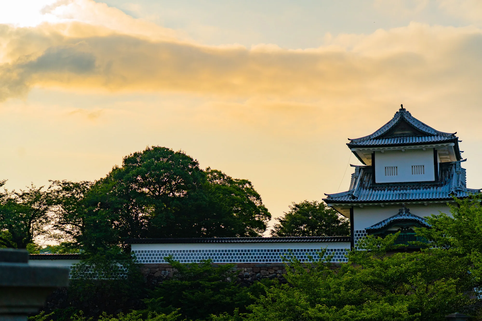 石川県金沢市にある金沢城を夕暮れ時に撮影した写真です。画面右側に城の櫓の一部が写っています。白壁に瓦屋根の美しい建物で、歴史を感じさせます。建物の周りには緑豊かな木々が生い茂り、自然に囲まれた城であることがわかります。空は夕焼けに染まり、オレンジ色や黄色のグラデーションが美しいです。雲も浮かんでおり、幻想的な雰囲気を醸し出しています。写真全体として、日本の歴史的な建造物と自然の美しさを組み合わせた、趣のある風景を描いています。静かで落ち着いた雰囲気が伝わってきます。
