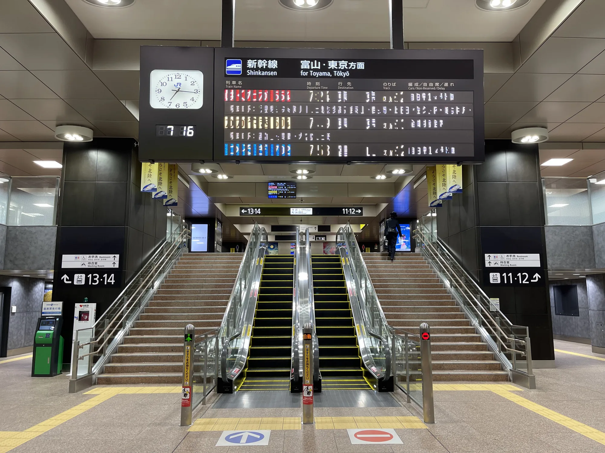 写真は金沢駅の新幹線乗り場を写したものです。正面には階段とエスカレーターが2基ずつ並んでいます。階段とエスカレーターの上には、新幹線の発車案内表示板が設置されており、時刻、列車名、行き先などが表示されています。表示板の左側にはアナログ時計が設置されており、現在時刻が7時16分であることを示しています。階段の左右には、プラットホームの番号を示す案内板（11-12番線、13-14番線）が見えます。プラットホームに続く通路の壁には「ようこそ北陸へ」という広告が掲示されています。床には黄色い誘導線と、進行方向を示す矢印マークや進入禁止マークが描かれています。全体的に明るく清潔感があり、整然とした雰囲気です。無機質でありながらも、どこか温かみを感じさせる日本の駅らしい光景が広がっています。