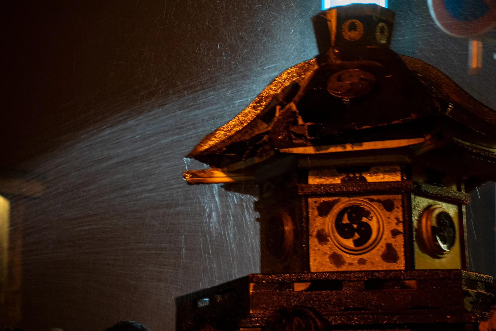 能登で撮影されたと思われる写真には、雨の中、祭りの山車の一部である提灯が写っています。夜であるため全体的に暗い印象ですが、提灯はオレンジ色の光を放っており、雨に濡れて光沢を帯びています。提灯の屋根には伝統的な模様が施され、四角い側面には「巴」の紋があしらわれています。提灯の周囲には雨が激しく降り注いでおり、光の筋となって写っていることから、雨の勢いが強く感じられます。画面奥は暗闇に包まれ、ぼんやりとした光が祭りの賑わいを暗示しているようです。雨に濡れた提灯の質感と、暗闇に浮かび上がる光のコントラストが印象的な一枚です。
