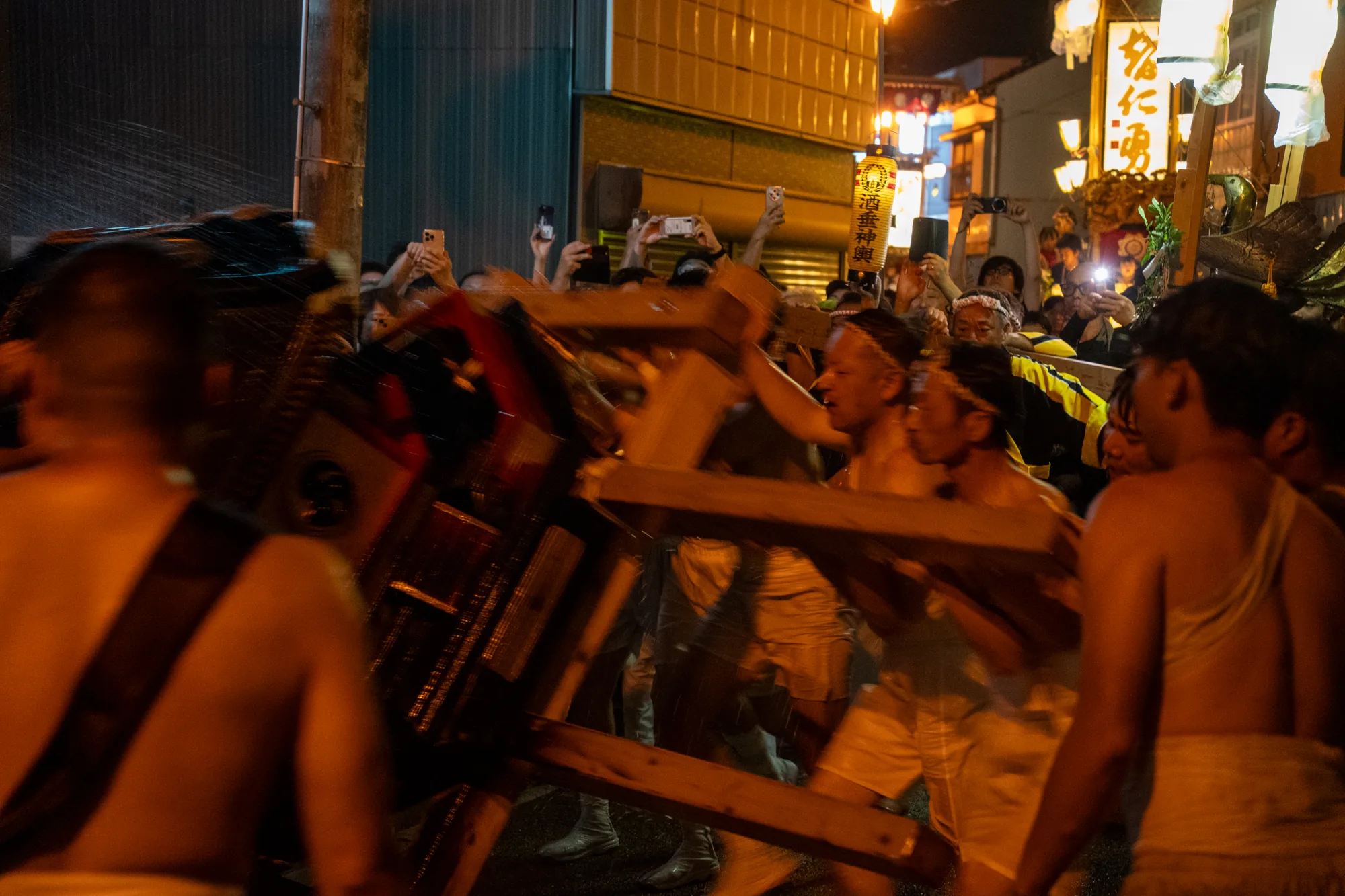 この写真は、日本の能登で撮影された夜の祭りの様子を捉えています。担ぎ手たちが木製の構造物を担いでおり、激しい動きと水しぶきから、勢いのある活気的な祭りであることが伺えます。担ぎ手たちは、ふんどし姿でハチマキを巻いており、伝統的な祭りの装いです。構造物の一部はぼやけており、動きの速さを強調しています。背景には、多くの人々がスマートフォンで撮影している様子が確認でき、祭りの賑わいを伝えています。また、灯籠や看板には日本語の文字が見られ、日本独特の祭りの雰囲気を醸し出しています。「酒菱神輿」と書かれた灯籠も確認できます。全体的に、熱気と興奮に満ちた、能登の伝統的な祭りの瞬間を切り取った一枚と言えるでしょう。