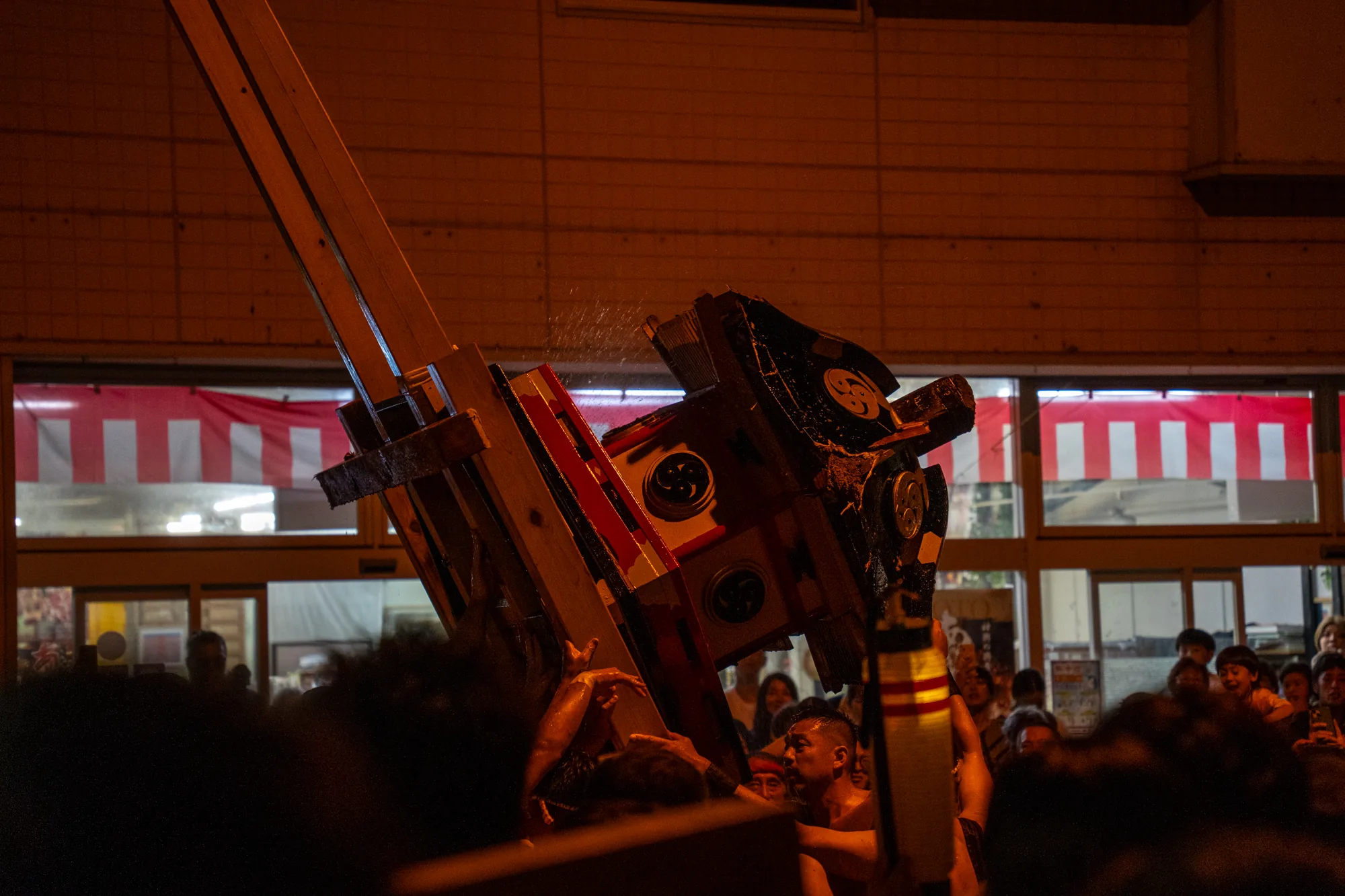 この写真は、日本の能登で撮影された祭りの様子を捉えています。夜間の撮影で、照明は控えめですが、祭りの熱気が伝わってきます。中心には、巨大な木製の構造物が、何人もの人々によって担ぎ上げられています。この構造物は、細部まで装飾が施されており、黒い背景に赤と白のアクセントが映えます。担ぎ手たちは上半身裸で、汗をかきながら力を込めています。彼らの表情は真剣そのもので、祭りに情熱を注いでいる様子がうかがえます。構造物からは水が飛び散っており、動きと活気が感じられます。背景には、商店らしき建物の窓が見え、赤と白のストライプ模様の布が飾られています。見物客も多く、祭りの様子を興奮気味に見守っています。全体的に、この写真は能登の祭りの力強さとエネルギーを伝える一枚です。