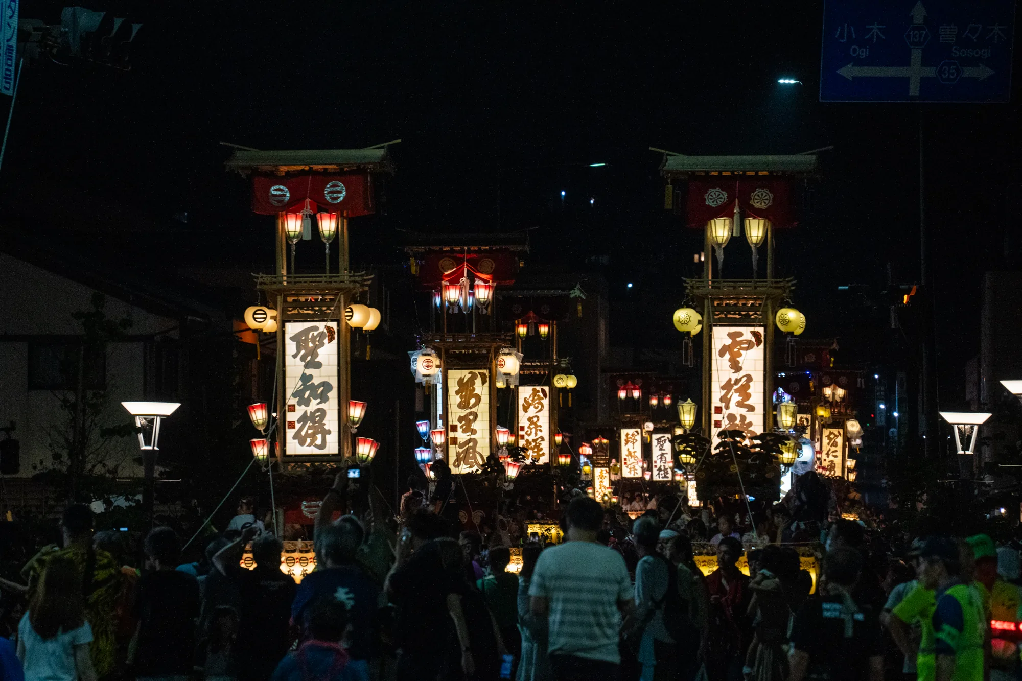 写真は日本の能登で撮影された夜祭りの様子を捉えています。暗闇の中に、複数の祭りの山車が立ち並び、それぞれに灯籠が飾られて幻想的な雰囲気を醸し出しています。山車の前には大勢の見物客が集まっており、祭りの賑わいが伝わってきます。山車の灯籠には漢字が書かれており、祭り特有の伝統的な雰囲気を強調しています。背景には道路標識が見え、「小木」「曽々木」といった地名が確認できます。写真全体は、夜の暗さと灯籠の光のコントラストが美しく、祭りの熱気と静けさが混ざり合った独特の雰囲気を表現しています。
