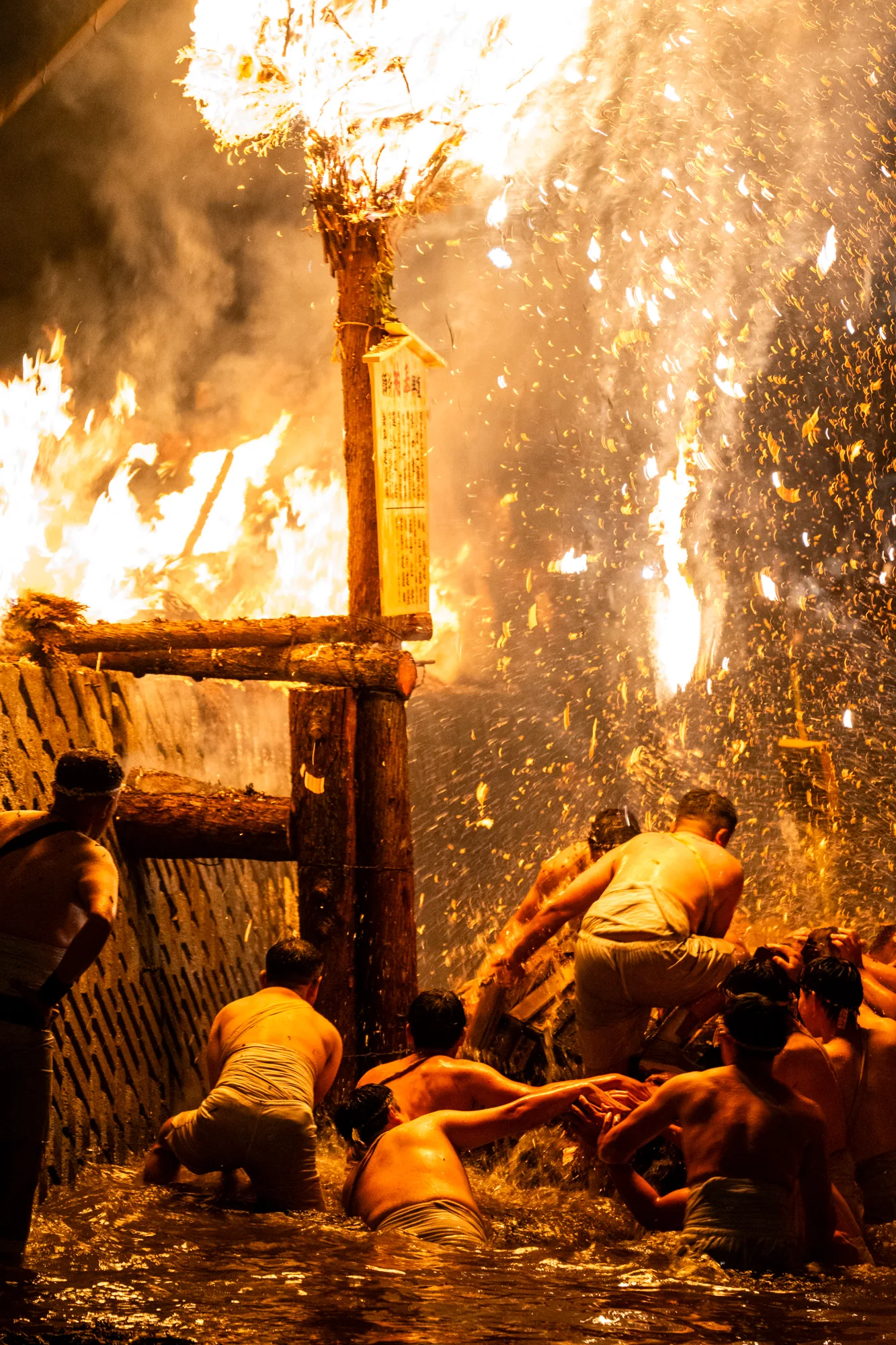 写真は日本の能登地方で撮影されたお祭りの様子を捉えています。激しく燃え盛る炎が画面全体を照らし出し、非常に熱狂的な雰囲気を醸し出しています。画面中央には高く組まれた木組みがあり、その上に積まれたものが激しく燃えています。木組みには札のようなものが取り付けられています。周囲には、ふんどし姿の男たちが水中に浸かりながら集まっており、火の勢いに耐えながら何かをしているようです。飛び散る火の粉が、その場の熱気と危険な雰囲気を伝えます。光と影のコントラストが非常に強く、祭りのエネルギーをダイナミックに表現しています。全体的に、能登地方の伝統的な火祭りの様子を力強く捉えた一枚です。
