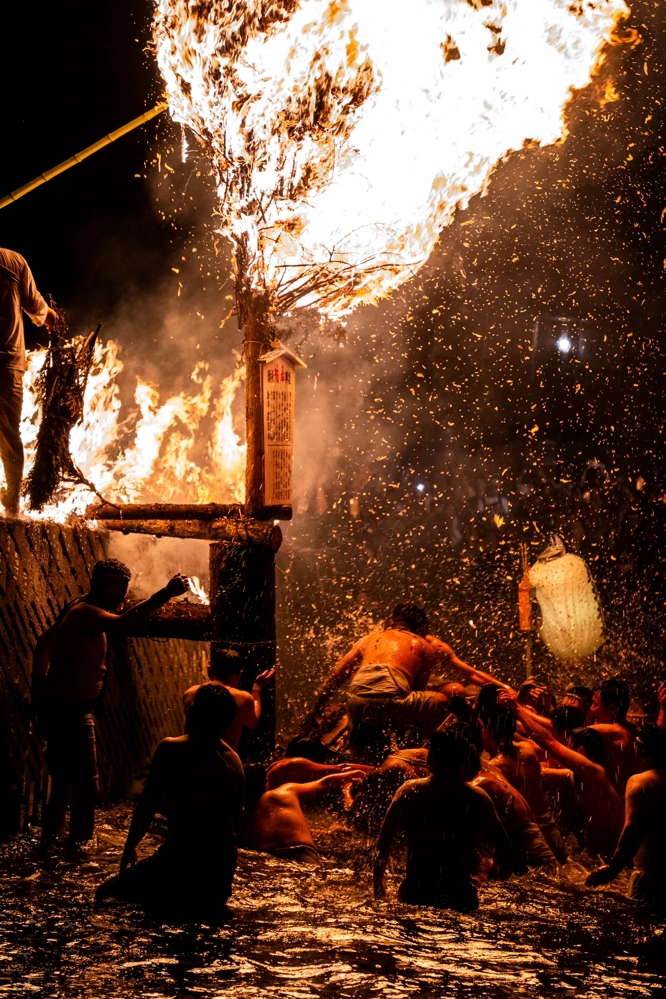 この写真は、日本の能登で行われている祭りの様子を捉えたものです。夜の暗闇の中、巨大な炎が激しく燃え上がり、その周りには上半身裸の男たちが集まっています。炎は、木や藁で組まれた巨大な構造物を燃やしており、火の粉が舞い上がり、幻想的な光景を作り出しています。男たちは水の中に浸かりながら、炎に向かって手を伸ばしたり、構造物を支えたりしており、祭りの熱気と興奮が伝わってきます。構造物には札のようなものも取り付けられており、宗教的な意味合いも感じられます。全体的に、力強く、エネルギーに満ち溢れた写真です。