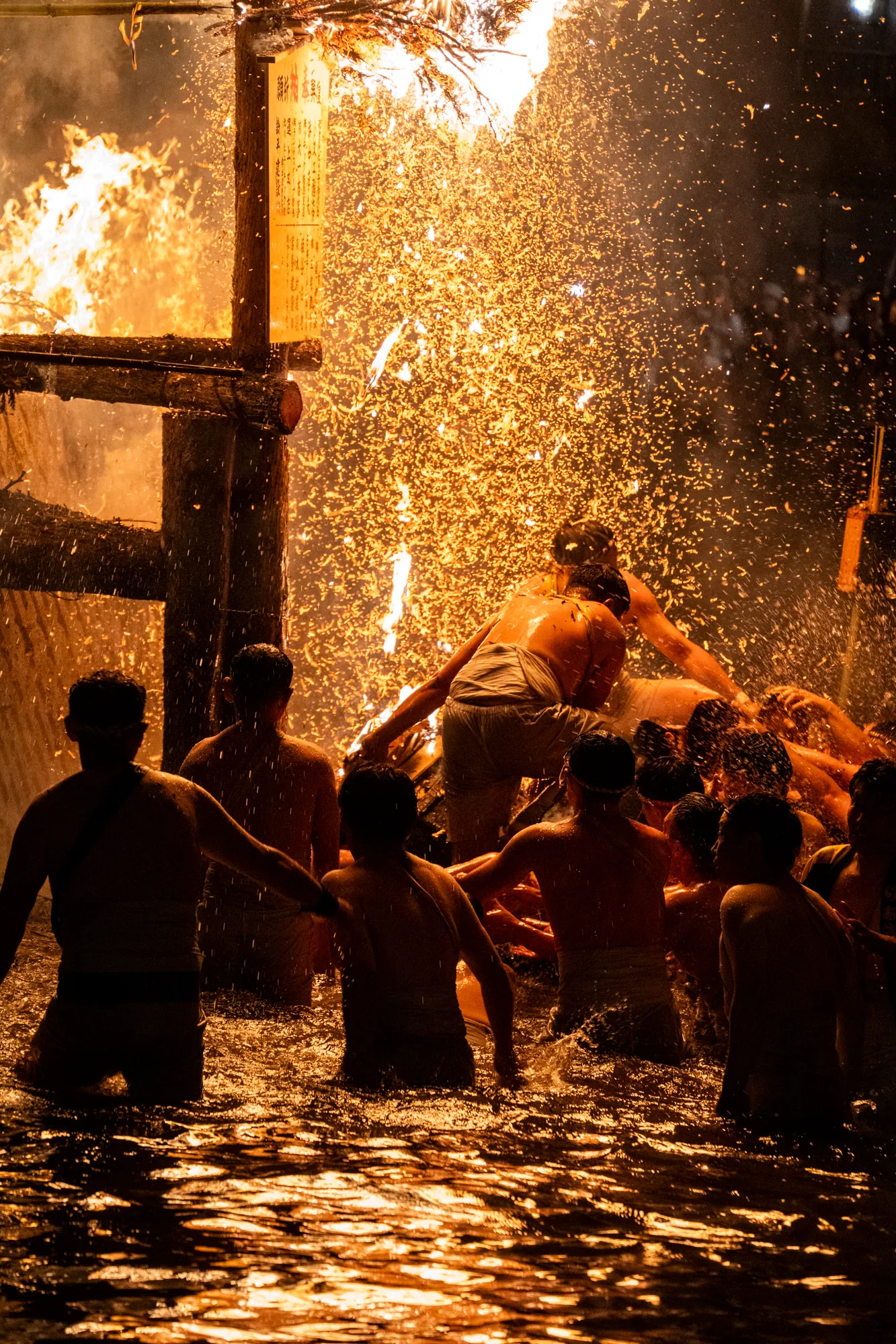 この写真は、日本の能登地方で撮影された火祭りの様子を捉えています。夜の暗闇の中、燃え盛る炎が強烈な光を放ち、周囲を照らしています。水の中に人々が集まり、炎の近くで何かを行っているようです。彼らは上半身裸で、腰に白い布を巻いているのが見えます。飛び散る火の粉が水面や人々の肌を照らし、躍動感と熱気を伝えています。炎の勢いと水しぶき、人々のシルエットが組み合わさり、幻想的で力強い雰囲気を醸し出しています。伝統的な祭りのエネルギーと、人々の熱気が感じられる一枚です。