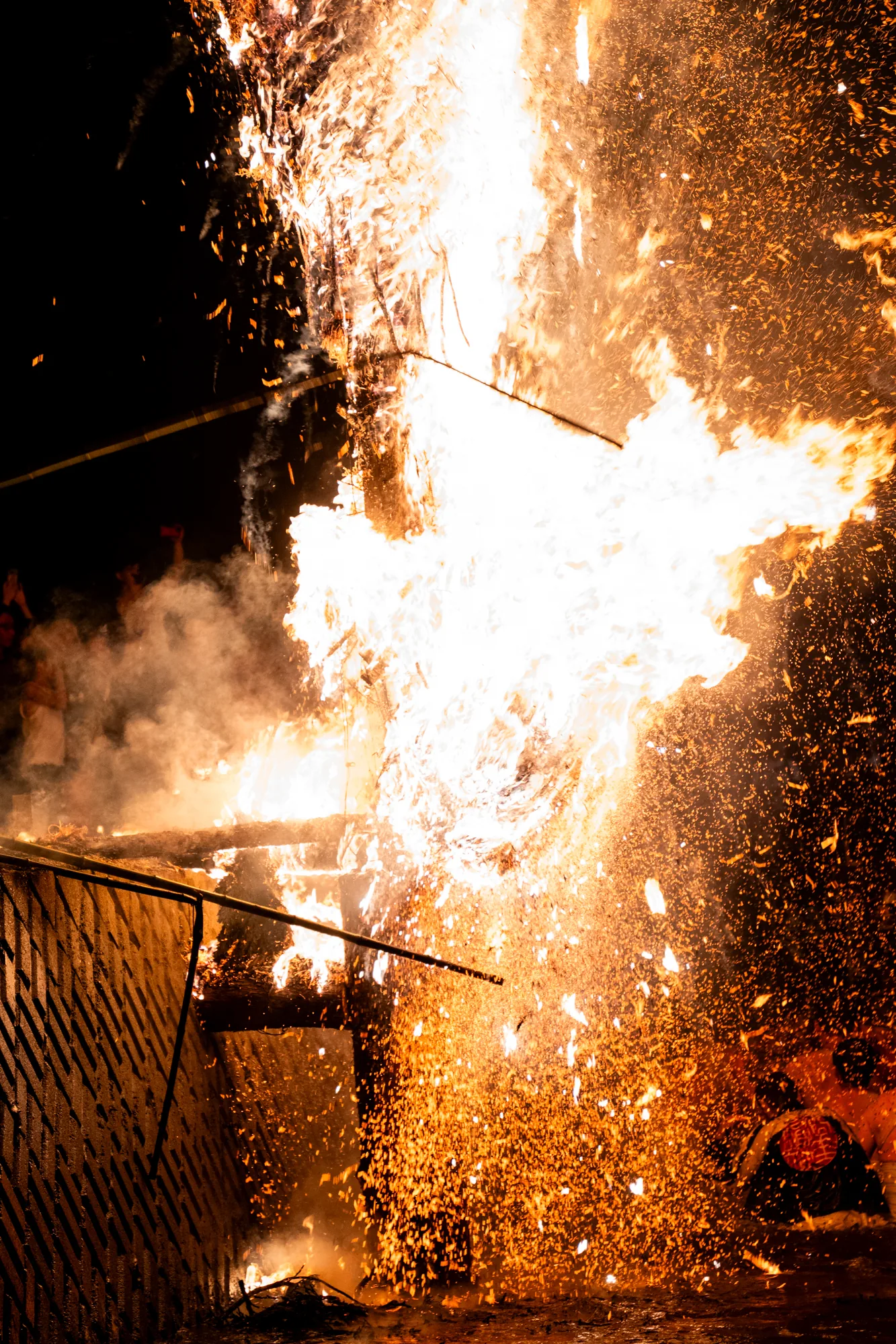 この写真は、日本の能登で撮影された炎上祭りの様子を捉えたものです。夜の闇を背景に、巨大な炎が激しく燃え盛っており、その周囲には無数の火の粉が飛び散っています。炎は、木材で組まれた構造物を焼き尽くしているようで、その形状が辛うじて見て取れます。構造物の手前には柵のようなものがあり、炎から観客を保護しているのかもしれません。写真の左側には、炎の熱気で揺らめく煙と、祭りを盛り上げる人々の姿がぼんやりと写っています。彼らは祭りの様子を写真に収めようとしているようです。全体的に、祭りの熱気とエネルギー、そして炎の壮大さを感じさせる一枚です。
