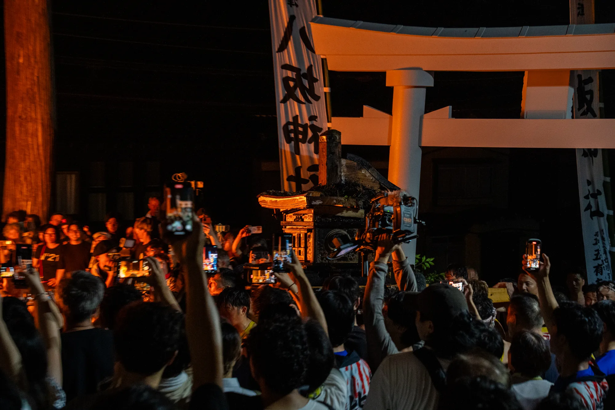 写真は、日本の能登で撮影されたと思われる、夜間の祭りの様子を捉えています。大勢の人々がスマートフォンを高く掲げ、何かの出来事を撮影しているようです。背景には、白く照らし出された鳥居と、「八坂神社」などの文字が書かれた幟が見えます。人々の視線の先には、神輿のようなものがあり、飾り付けが施されているようです。全体的に暗い環境ですが、神輿や鳥居、人々のスマートフォンから発せられる光が、祭りの熱気を伝えています。祭りの高揚感と、それを記録しようとする人々の様子が印象的な一枚です。