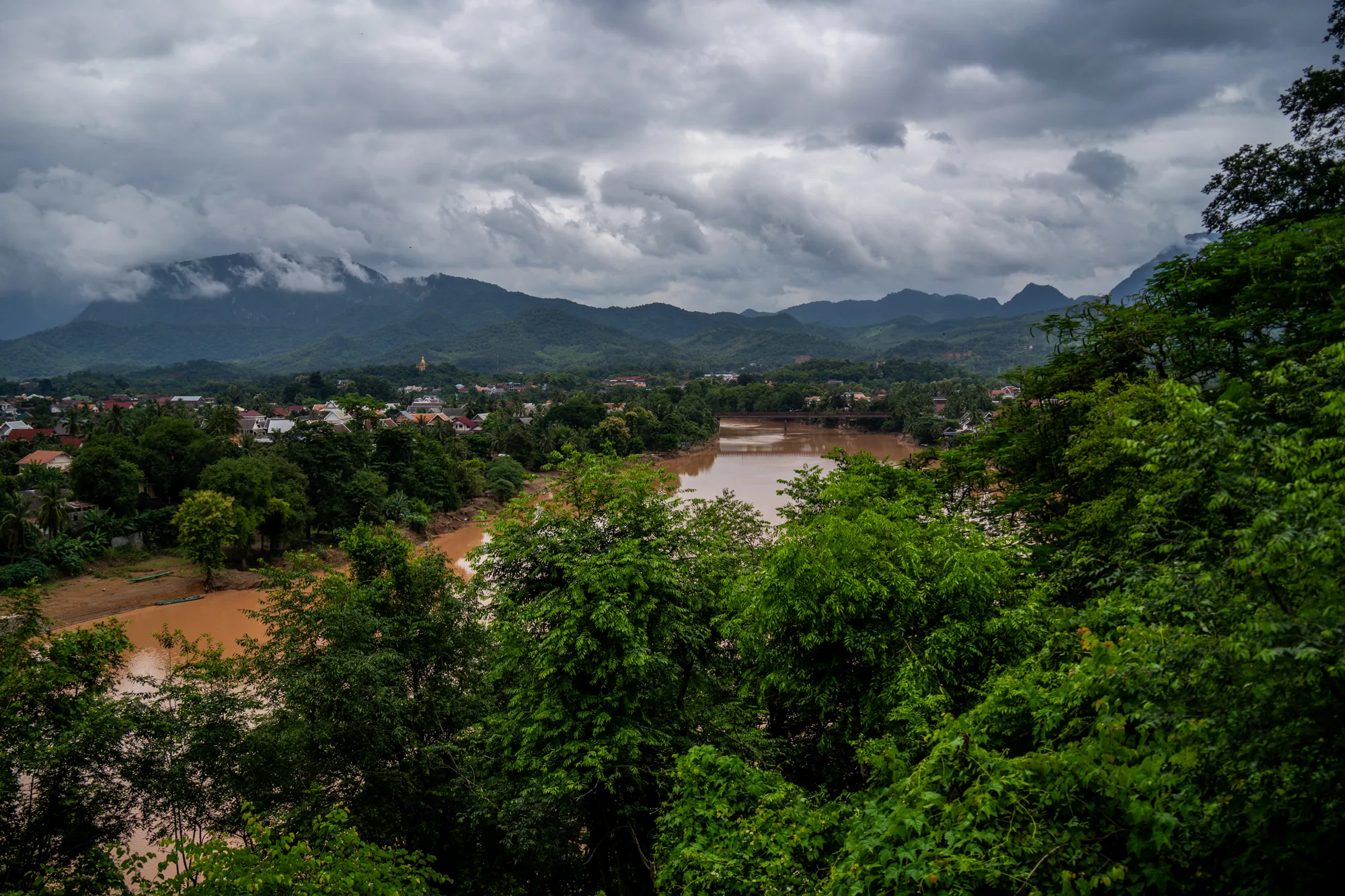 ラオスのルアンパバーンで撮影された写真です。画面全体は曇り空に覆われており、今にも雨が降り出しそうな重たい空気が漂っています。手前には緑豊かな木々が生い茂り、画面の半分ほどを覆っています。その奥には茶色く濁ったメコン川が流れ、川には橋が架かっています。川の両岸には家々が建ち並び、その先には緑の山々が連なっています。山頂付近は雲に覆われており、幻想的な雰囲気を醸し出しています。全体的に落ち着いた色調で、静かで穏やかな風景が広がっています。