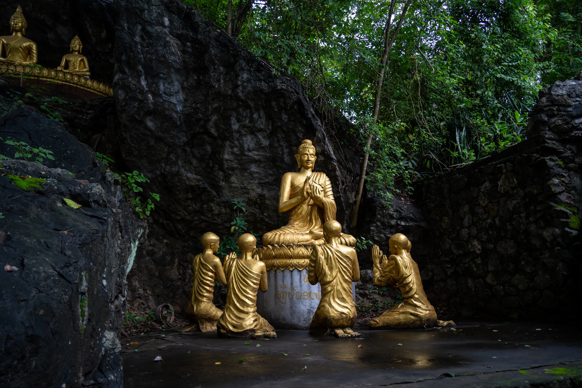 この写真はラオスのルアンパバーンで撮影されたもので、金色の仏像群が写っています。写真の中央には、座禅を組んだ大きな仏像があり、その周りには跪いて祈りを捧げる複数の僧侶の像があります。これらの像はすべて金色で、周囲の緑豊かな自然と対照的です。背景には岩肌が見え、その上には緑が生い茂っています。場所は神聖な場所であると思われます。地面は湿っていて、しっとりとした雰囲気が感じられます。全体的に、静寂と敬虔さが漂う写真です。