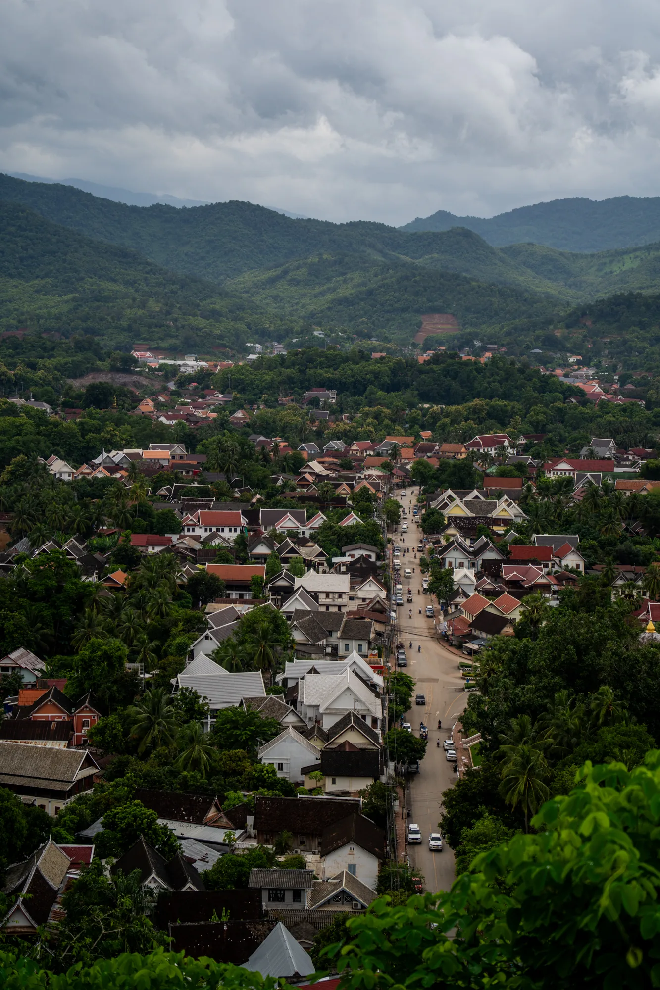 ラオスのルアンパバーンで撮影された写真です。高台から街全体を見下ろすように撮影されており、画面奥には緑豊かな山々が連なっています。空は曇り空で、しっとりとした空気が感じられます。手前の緑の木々をフレームにして、街並みが広がっています。家々の屋根は茶色や白、グレーなど様々な色合いで、木々に囲まれているのが印象的です。一本の道路が街の中心を貫いており、車やバイクが走行しているのが見えます。静かで落ち着いた雰囲気の、ルアンパバーンの美しい景観を捉えた一枚です。