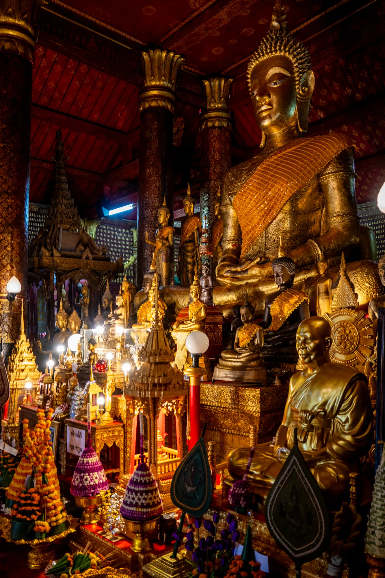 ラオスのルアンパバーンにある寺院で撮影された写真です。室内には、大小様々な黄金色の仏像が所狭しと並べられています。特に目を引くのは、画面右上にそびえ立つ巨大な仏像で、光沢のある金色に輝き、威厳を放っています。他の仏像も同様に金色の輝きを放ち、それぞれが異なるポーズや表情をしています。 仏像の手前には、お供え物と思われる花や装飾品が並べられ、華やかさを添えています。背景には、赤色の天井や金色の柱が見え、寺院の荘厳な雰囲気を強調しています。室内は薄暗く、仏像や装飾品がライトアップされていることで、神聖な空間が演出されています。全体的に豪華絢爛でありながら、静寂と神秘に満ちた印象を受ける写真です。