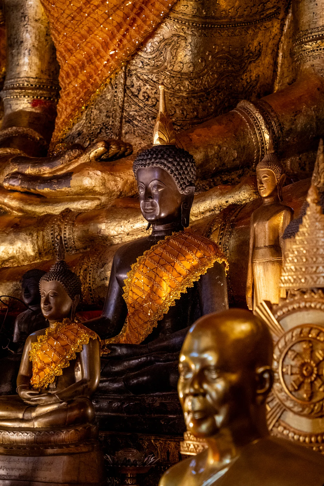 この写真はラオスのルアンパバーンで撮影されたものと思われます。画面全体に複数の仏像が写っており、荘厳な雰囲気を醸し出しています。背景には巨大な金色の仏像の一部が見え、その手や足には精緻な模様が施されています。  前景にはいくつかの小さな仏像が配置されており、中央の像は黒色の肌を持ち、鮮やかなオレンジ色の袈裟を身につけています。他の像は金色に輝き、それぞれ異なる表情や姿勢をしています。特に手前のぼやけた像は、頭部が強調されており、その表情を想像させる余地を残しています。  写真全体の色調は金色と暗色が組み合わさり、神聖で神秘的な雰囲気を強調しています。光の当たり具合によって仏像の表面が輝き、その存在感を際立たせています。細部まで丁寧に作り込まれた仏像の装飾や表情は、ラオスの仏教文化の深さを感じさせます。