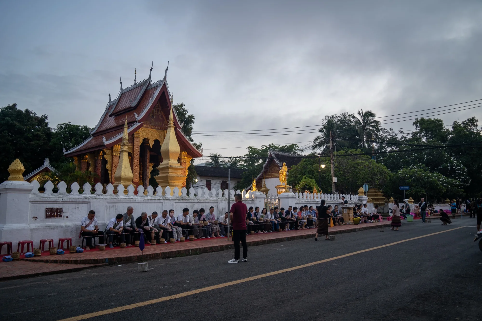 写真はラオスのルアンパバーンで撮影された托鉢の様子を捉えたものです。早朝、薄暗い空の下、道路沿いに人々が腰掛けており、僧侶への供え物を手に持っています。手前には、赤や青の小さな椅子と、お供え物を入れると思われる器が並んでいます。道路を挟んで奥には、豪華な装飾が施された寺院が見えます。寺院は赤茶色の屋根を持ち、金色の装飾が輝いています。白い塀が寺院を囲み、神聖な雰囲気を醸し出しています。空は曇り空で、静寂な空気感が漂っています。人々の表情は穏やかで、敬虔な気持ちが伝わってきます。ルアンパバーンの伝統的な文化と信仰を感じさせる一枚です。