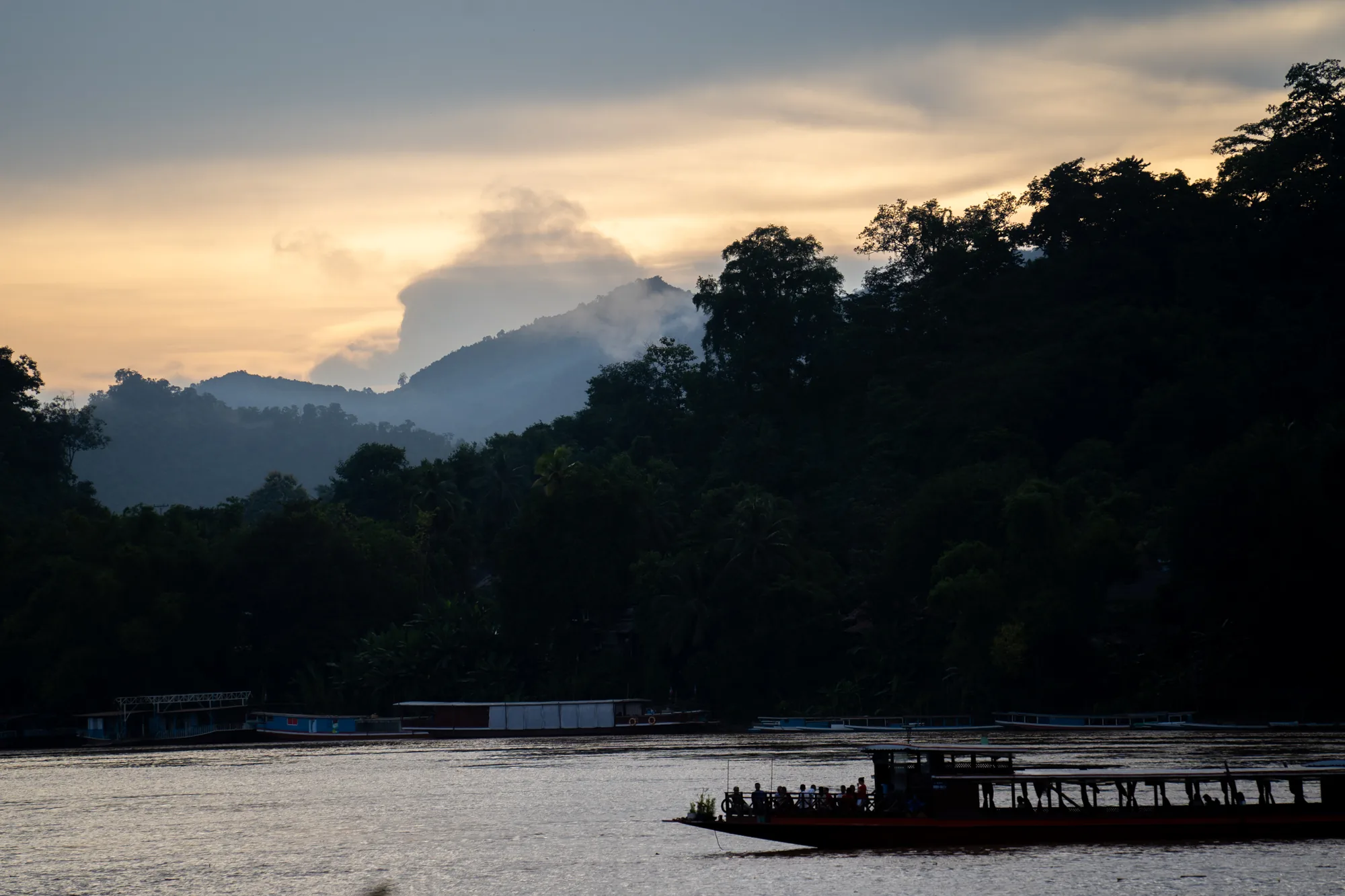 ラオスのルアンパバーンで撮影された写真です。夕暮れ時、川面に夕日の光が反射し、穏やかな雰囲気を醸し出しています。手前には人が乗ったボートが写っており、シルエットになっています。奥には、いくつかの船が停泊しています。背景には、木々が生い茂る山々が連なり、一部は靄に包まれています。空は、夕焼けのオレンジ色と雲の灰色が混ざり合い、幻想的な景色を作り出しています。全体的に、静かで落ち着いた印象を与える写真です。