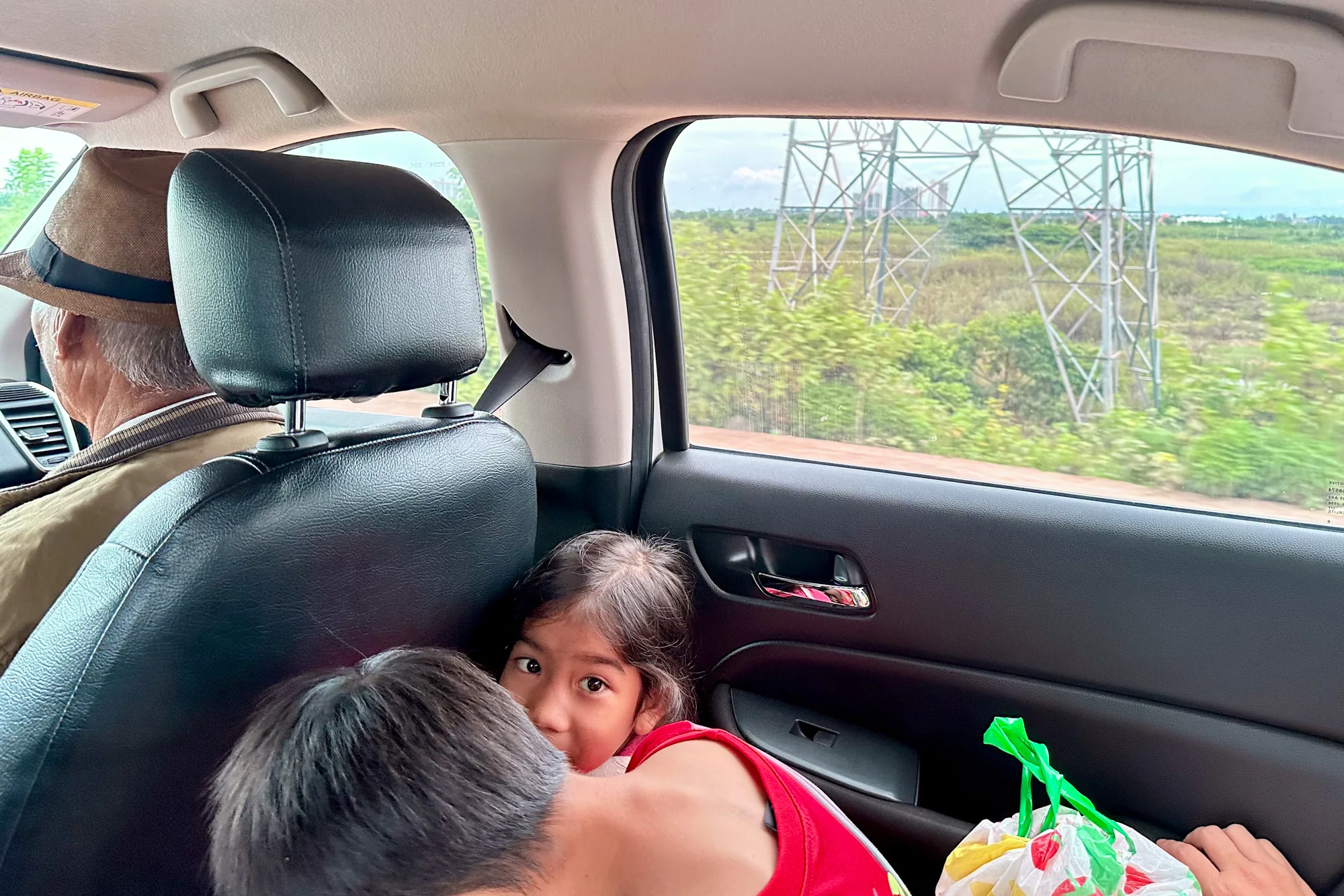 ラオスのビエンチャンで撮影された写真です。車の後部座席にいる家族の様子が写っています。助手席には帽子を被った高齢の男性が座っており、後部座席には幼い女の子が、身を乗り出してカメラの方を見ています。おそらく兄弟であろう少年が女の子の隣にいます。窓の外には、緑豊かな風景と鉄塔が見えます。車内のドアには緑の持ち手がついた買い物袋が置かれています。全体的に温かく、旅の途中の家族の親密な瞬間を捉えた写真です。