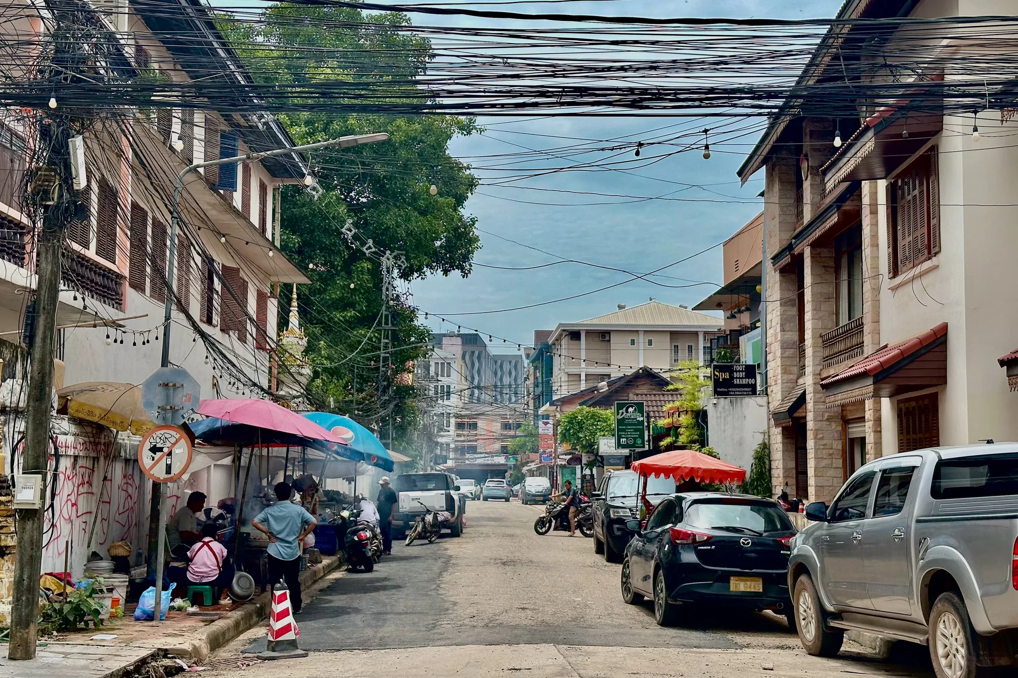 写真はラオスのヴィエンチャンで撮影された街並みを捉えています。通りは舗装されており、両側には建物が立ち並び、多くの電線が空を覆っています。左側の歩道には屋台が出ており、人々が座って食事をしている様子が見られます。屋台の近くには、路上喫煙禁止の標識が立っています。通りには車やバイクが駐車しており、交通量はそれほど多くありません。写真全体から、ヴィエンチャンの穏やかでローカルな雰囲気が伝わってきます。建物の様式は西洋の様式に影響されているようです。