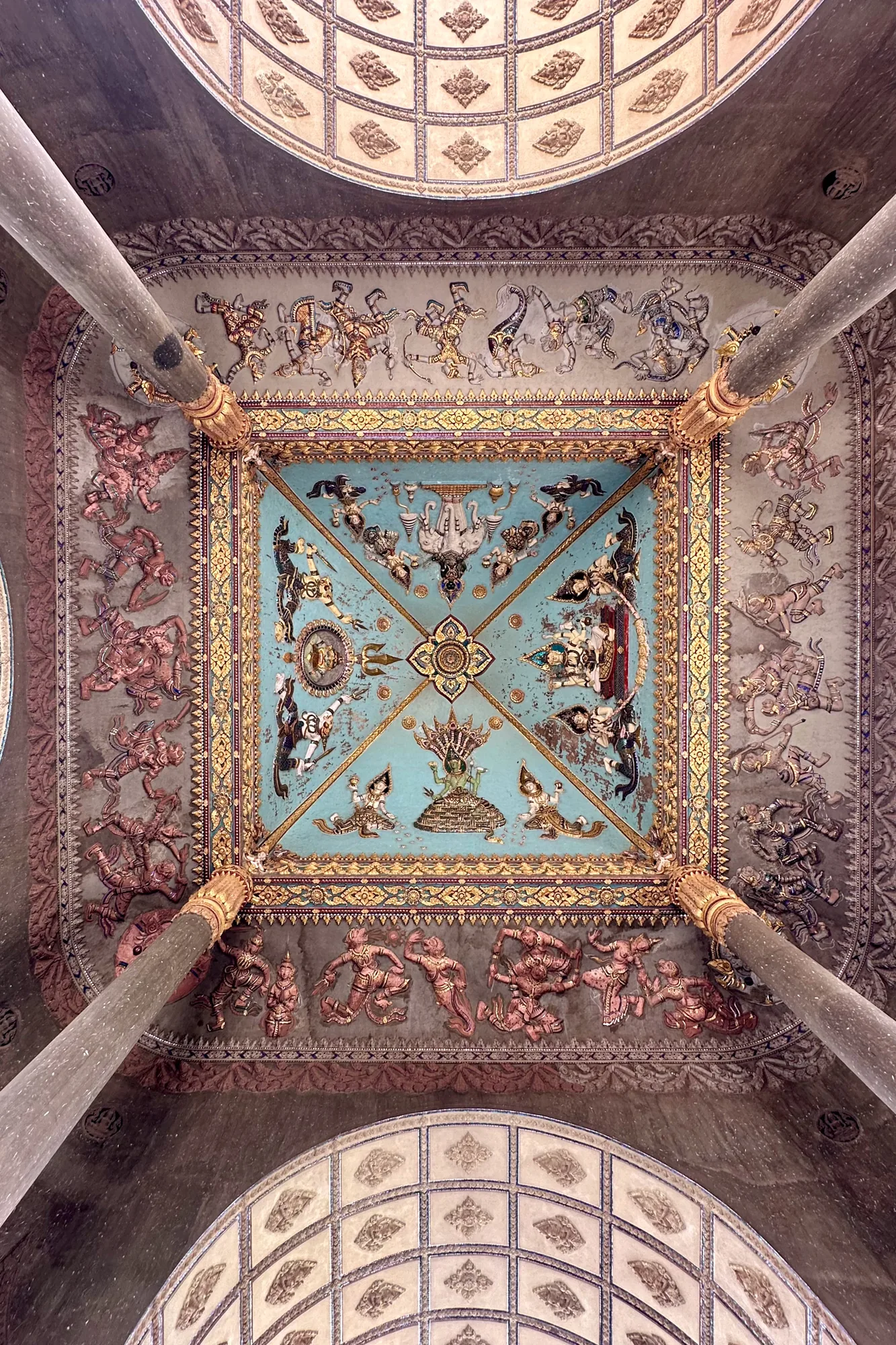 ラオスのビエンチャンで撮影された写真には、伝統的な装飾が施された建物の内部、特に天井が写っています。天井は正方形で、中央部分は水色に塗られ、金色の装飾が施されています。その中央には仏像や神話的な生き物、幾何学模様が描かれており、仏教的なモチーフが見られます。天井の四隅には、物語の一場面を描いたと思われる人物像が彫刻されています。これらの人物像は、踊っているように見えたり、武器を持っているように見えたりするものもあります。天井を支える柱が4本見えており、それぞれが金色で装飾されています。全体的に、歴史的、文化的価値の高い場所であると感じさせる雰囲気です。