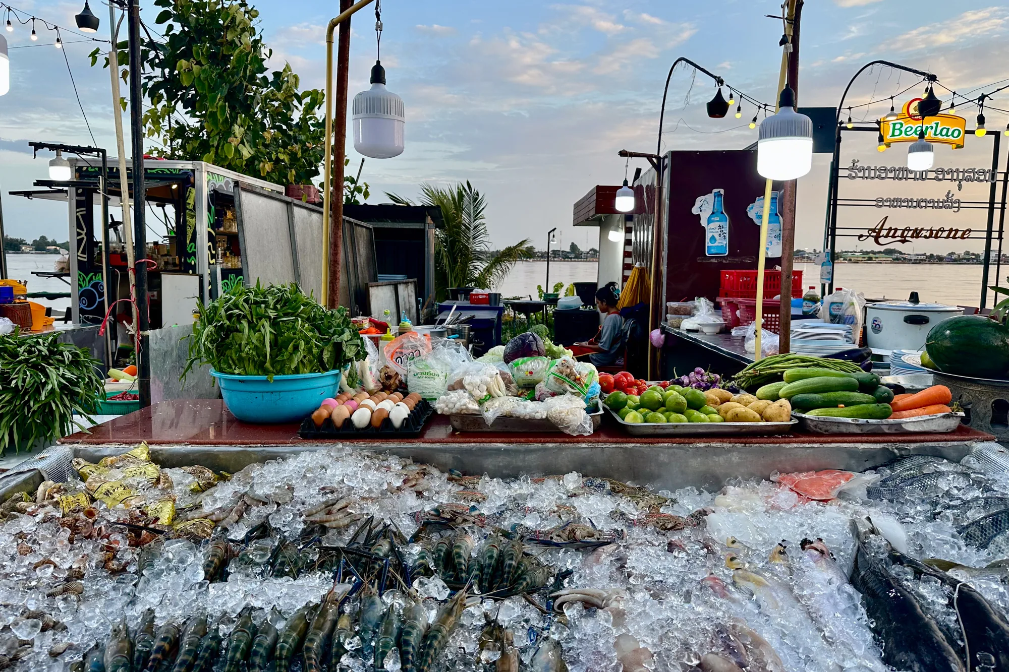 この写真は、ラオスのビエンチャンで撮影された市場の風景を捉えています。手前には、氷の上に新鮮な魚介類が並べられた露店が写っています。エビ、カニ、魚などが豊富に並べられ、活気ある市場の様子が伝わってきます。露店の奥には、さまざまな種類の野菜や卵が置かれています。さらに奥には、調理場らしきスペースがあり、人が作業をしている様子も見えます。背景にはメコン川が流れ、夕暮れ時の空が穏やかな雰囲気を醸し出しています。画面右奥には「Beerlao」と書かれた看板が見え、地元ビールが提供されていることも伺えます。全体的に、新鮮な食材と活気ある雰囲気が感じられる写真です。
