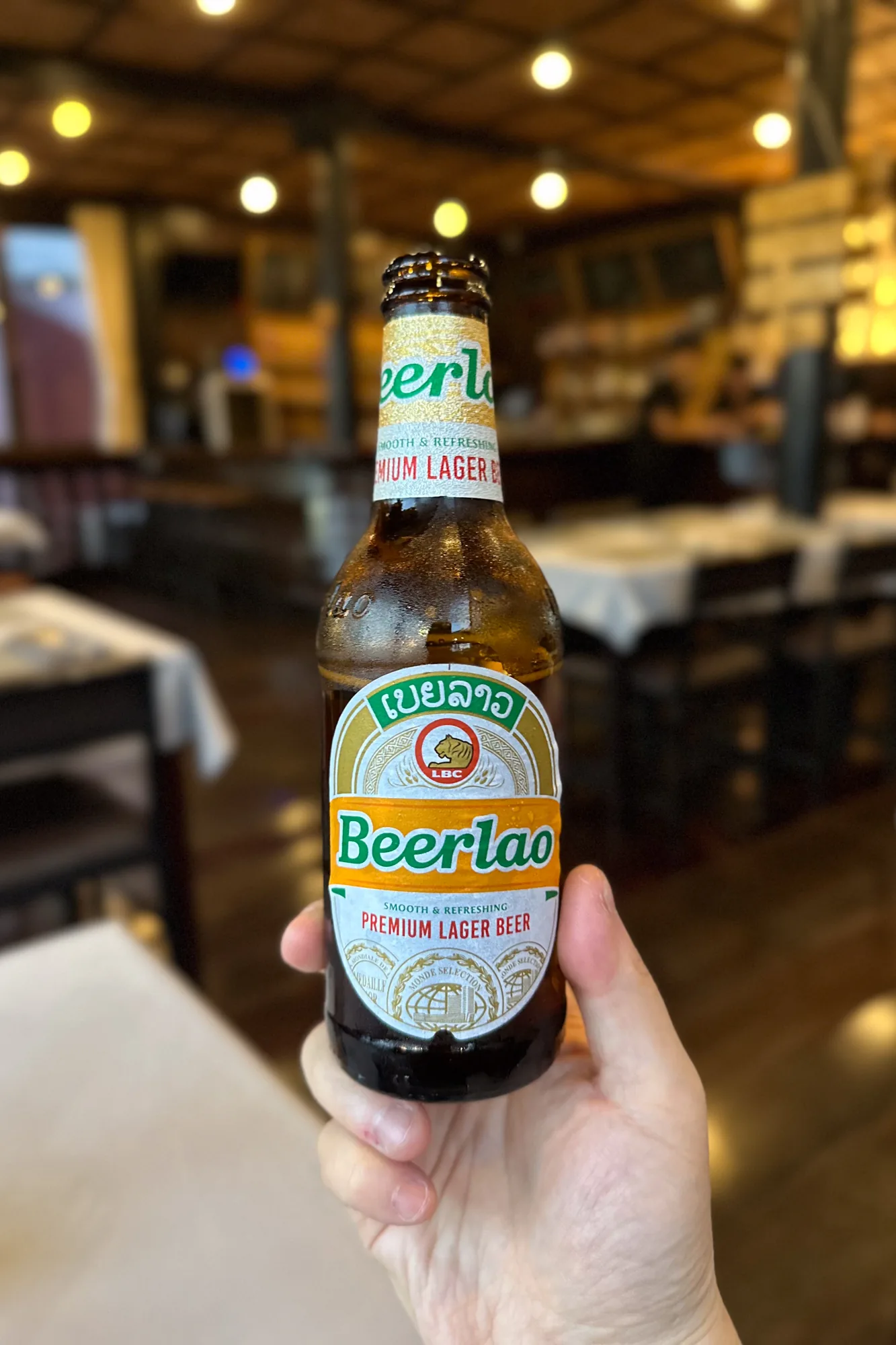 写真には、ラオスのビエンチャンにあるレストランと思われる場所で、ビアラオのボトルが写っています。手で持たれているビアラオのボトルには水滴が付いており、冷えていることがわかります。ラベルには「Beerlao」と大きく書かれており、その上にラオス語で「ເບຍລາວ」と書かれたロゴ、そしてLBCと虎の絵が描かれています。背景にはテーブルと椅子がぼやけて写っており、レストランの雰囲気が伝わってきます。天井には丸い照明がいくつか見えます。全体的にカジュアルな雰囲気で、旅行者が現地のビールを楽しんでいる様子が想像できます。