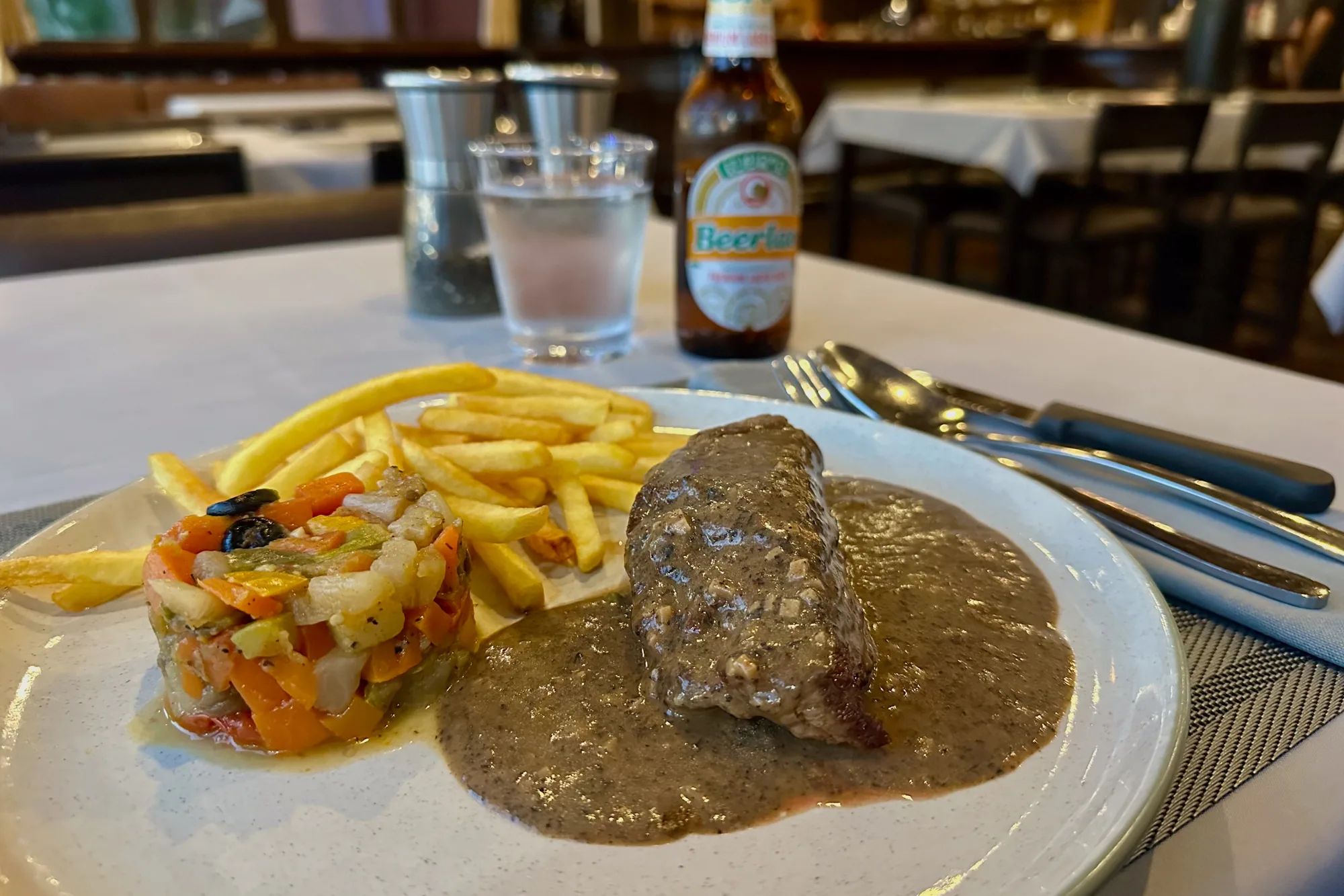 ラオスのビエンチャンで撮影された写真です。レストランのテーブルの上に、ステーキ、フライドポテト、野菜の重ね焼きが盛り付けられたお皿があります。ステーキは茶色いソースで覆われており、フライドポテトは黄金色でカリッとしていそうです。野菜の重ね焼きは、様々な色が鮮やかで食欲をそそります。背景には、水が入ったグラス、塩胡椒入れ、ビアラオのビール瓶が見えます。テーブルクロスは白く、カトラリーが丁寧に並べられています。レストランの落ち着いた雰囲気と、料理の美味しさが伝わってくる一枚です。