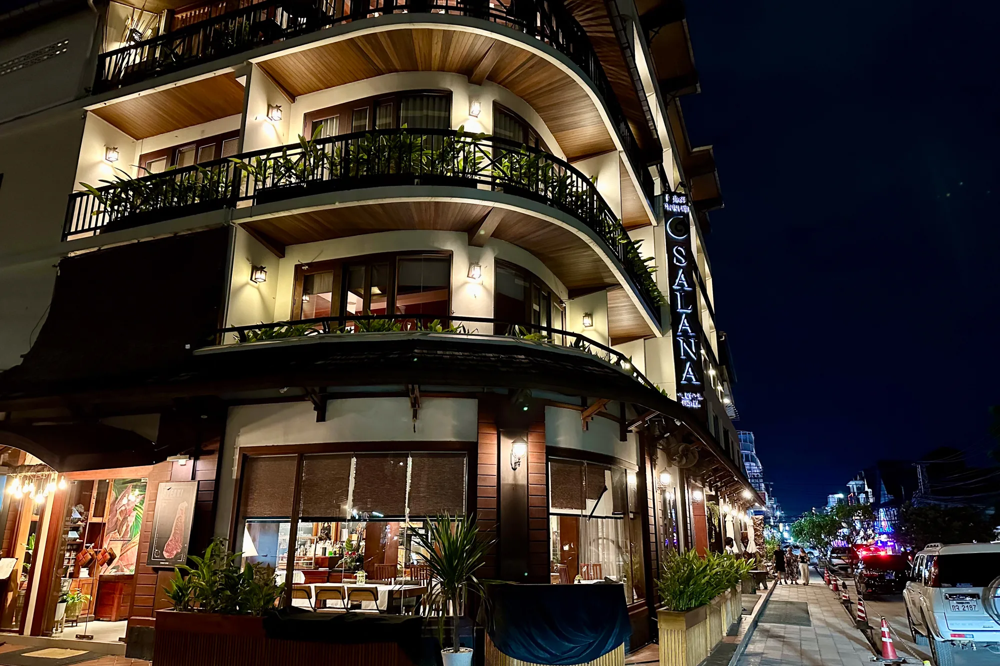 これはラオスのヴィエンチャンで撮影された夜間の写真です。写真には「SALANA」と書かれたホテルの一部が写っています。ホテルの建物は白を基調としており、木製の天井とバルコニーが特徴的です。各階のバルコニーには植物が飾られており、温かみのある照明が建物全体を照らしています。  ホテルの1階にはレストランがあり、窓からはテーブルや椅子がいくつか見えます。レストランの窓にはブラインドが設置されています。  ホテルの前の通りには、歩行者や車が行き交っています。街灯が灯り、夜の街並みを照らしています。空は濃い青色で、一部に雲が見えます。全体的に、写真からはヴィエンチャンの落ち着いた夜の雰囲気が伝わってきます。