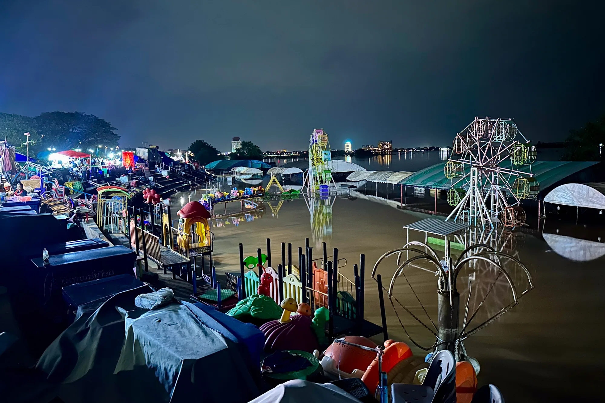 ラオスのビエンチャンの夜に撮影された写真です。広範囲に渡り水没しており、遊園地のような場所が水に浸かっている様子が写っています。手前にはテーブルや椅子が覆いをかぶせられた状態で置かれており、奥にはカラフルな遊具や乗り物が見えます。特に目立つのは、複数の観覧車で、ライトアップされて水面に反射し、幻想的な光景を作り出しています。遠景には建物の灯りも見え、都市の夜景が広がっています。全体的に暗く、水面が光を反射していることで、不気味でありながらもどこか美しい雰囲気を醸し出しています。水害の深刻さを伝えるとともに、人々の生活の痕跡が感じられる一枚です。
