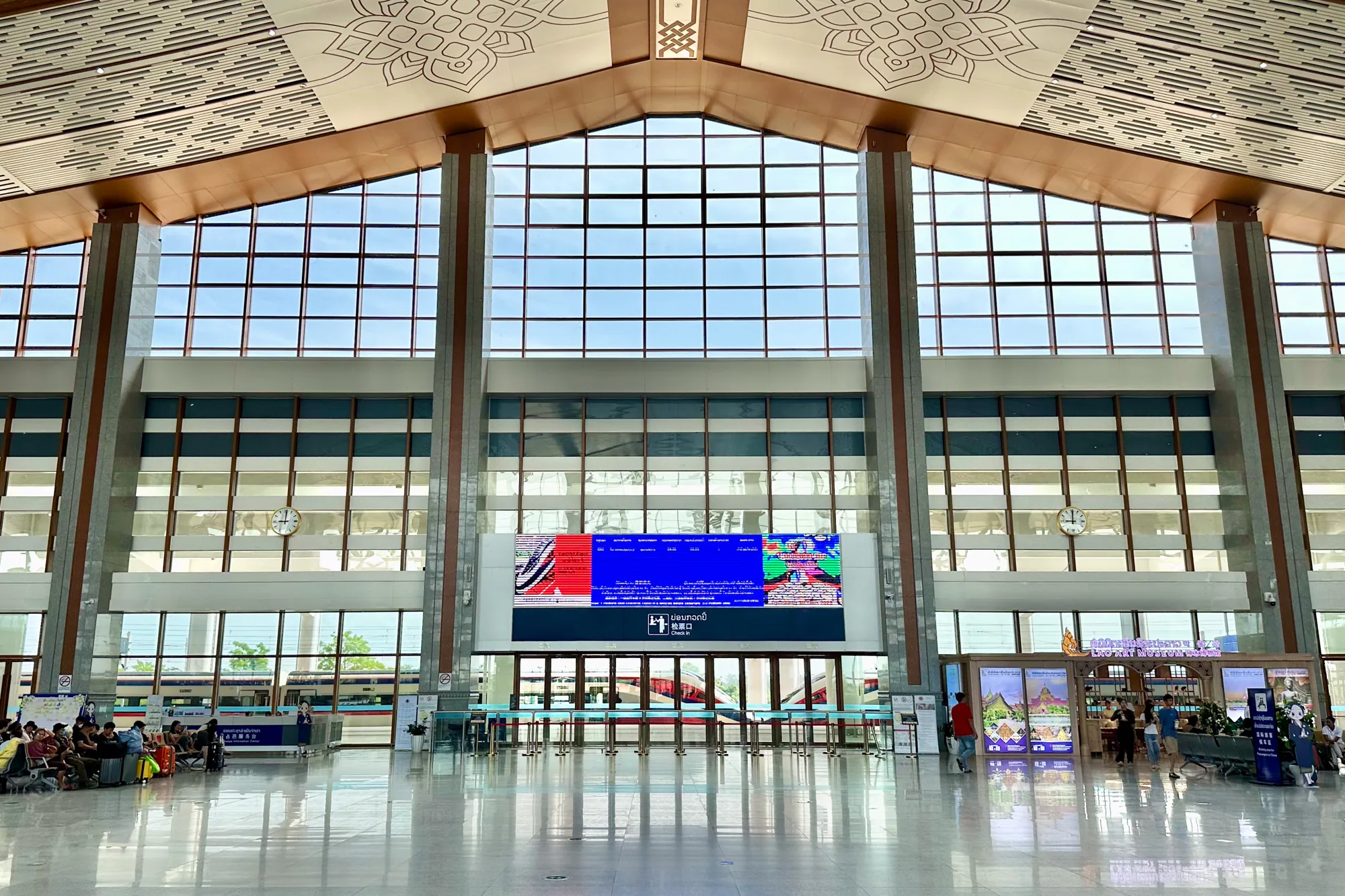 写真はラオスのビエンチャンにある駅の構内を捉えています。高い天井には幾何学的な模様が描かれ、大きな窓からは明るい光が差し込んでいます。窓の外には列車が見え、構内には待合スペース、案内カウンター、デジタルサイネージ、チェックインカウンターなどが確認できます。床は磨き上げられたタイル張りで、光を反射して明るい印象を与えています。待合スペースには旅行者が座って待機しており、構内には人々が歩いています。全体的に、広々として清潔感のある、モダンな駅の雰囲気です。