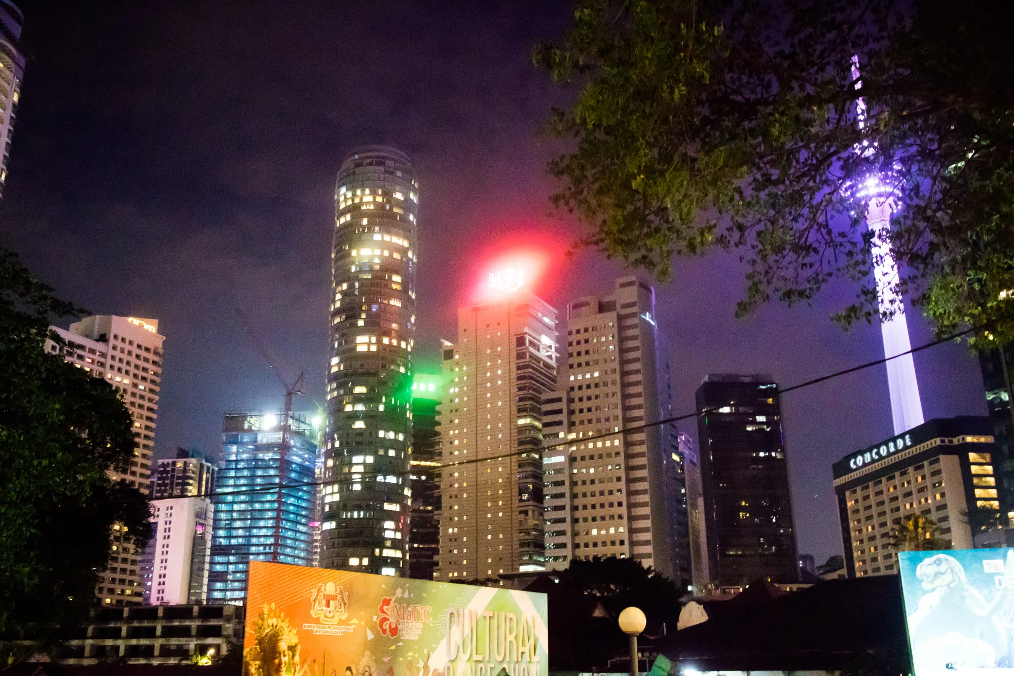 写真はマレーシアのクアラルンプールで撮影された夜景です。画面奥には高層ビル群が立ち並び、それぞれの窓から明かりが漏れています。特に目立つのは、円筒形の高層ビルで、多くの窓が光を放ち、その独特の形状を際立たせています。別のビルには、屋上に赤いライトが灯されており、その存在を際立たせています。遠くには、クアラルンプールタワーが紫色にライトアップされ、都市のランドマークとして輝いています。写真手前には街路樹の緑が一部写り込み、夜空とのコントラストを生み出しています。また、看板の一部が写っており、都市の活気と文化的な側面を垣間見ることができます。全体的に、夜のクアラルンプールが持つ都会的で華やかな雰囲気が伝わってくる一枚です。