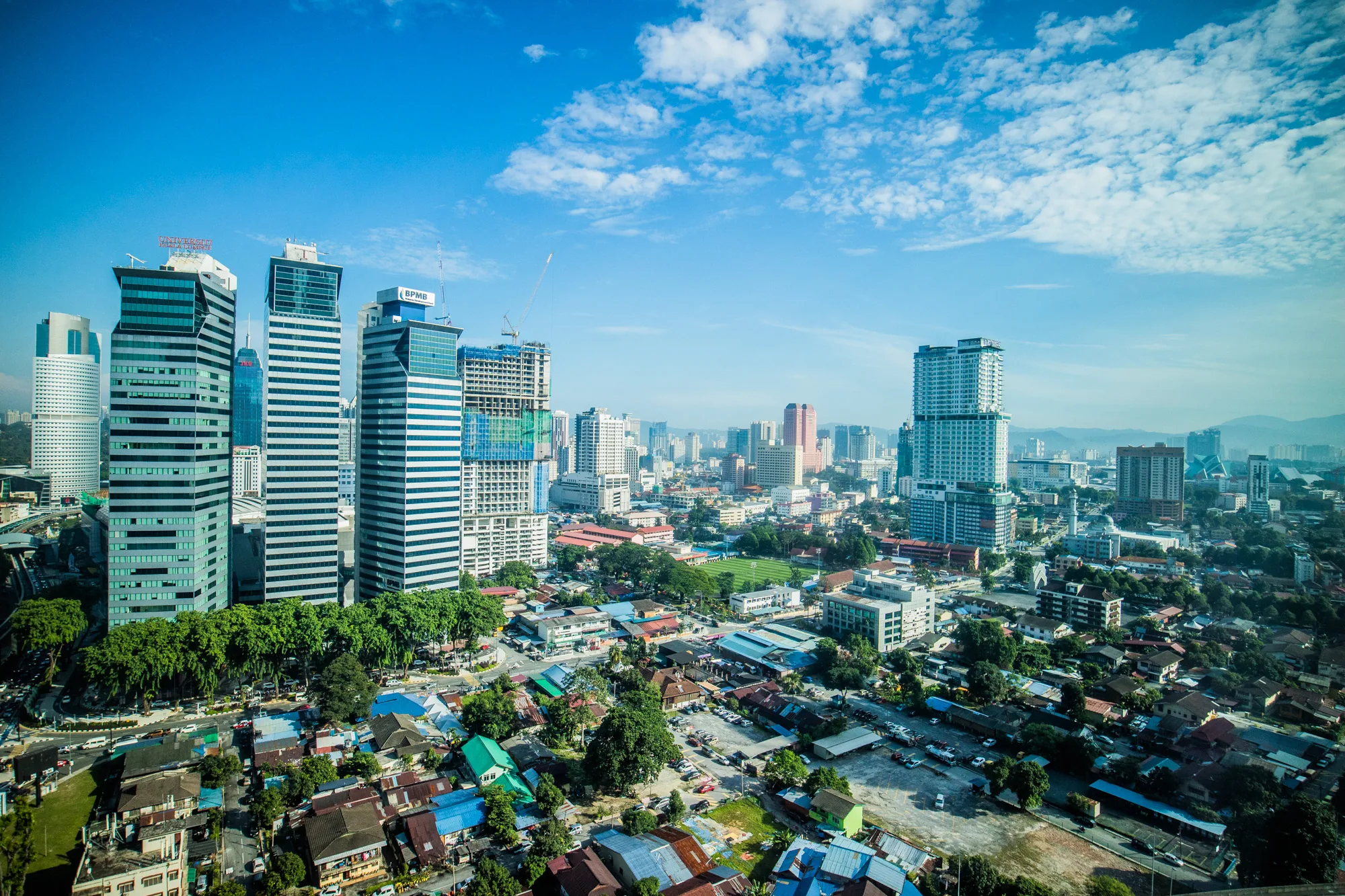 写真はマレーシアのクアラルンプールで撮影されたもので、高所から街並みを捉えています。手前には、様々な色の屋根を持つ住宅が密集して建ち並び、緑豊かな木々が点在しています。住宅街の奥には、近代的な高層ビル群がそびえ立っており、建設中の建物も見られます。空は青く、白い雲が浮かんでおり、街全体を明るく照らしています。写真全体から、活気があり、発展を続ける都市の様子が伝わってきます。遠くには山々がかすかに見え、都市と自然が調和している様子もうかがえます。