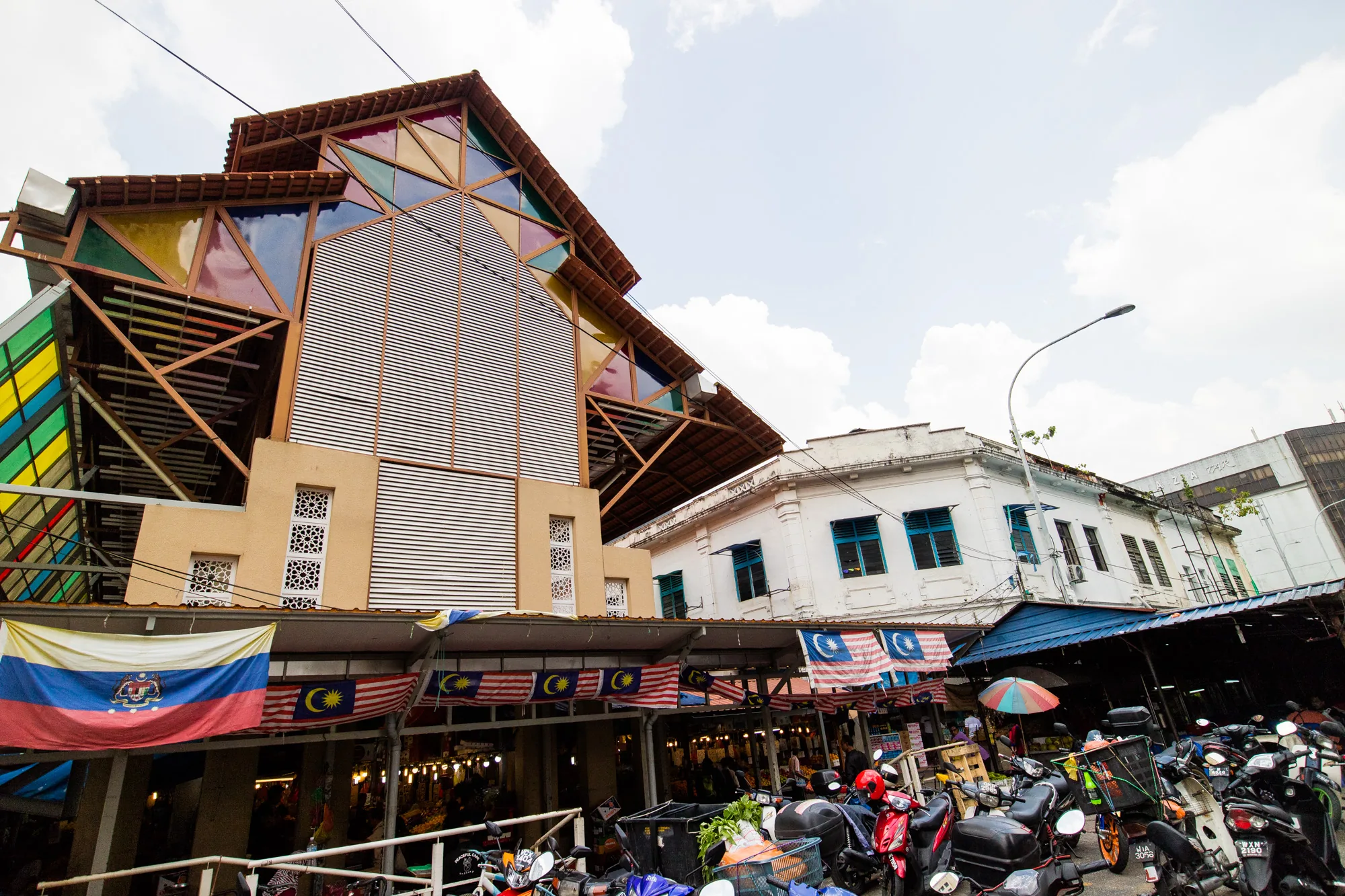 写真はマレーシアのクアラルンプールで撮影されたもので、市場の様子を捉えています。手前には多くのバイクが停められており、活気のある雰囲気を感じさせます。市場の屋根には、マレーシアの国旗が飾られています。背景には、独特なデザインの建物が写っており、カラフルなステンドグラスのような装飾が目を引きます。また、白い壁に青い窓枠の建物も見え、歴史を感じさせる佇まいです。空には白い雲が浮かび、明るい日中の様子が伺えます。全体的に、都市の喧騒と多様な文化が混ざり合ったような、クアラルンプールの魅力が伝わってくる写真です。