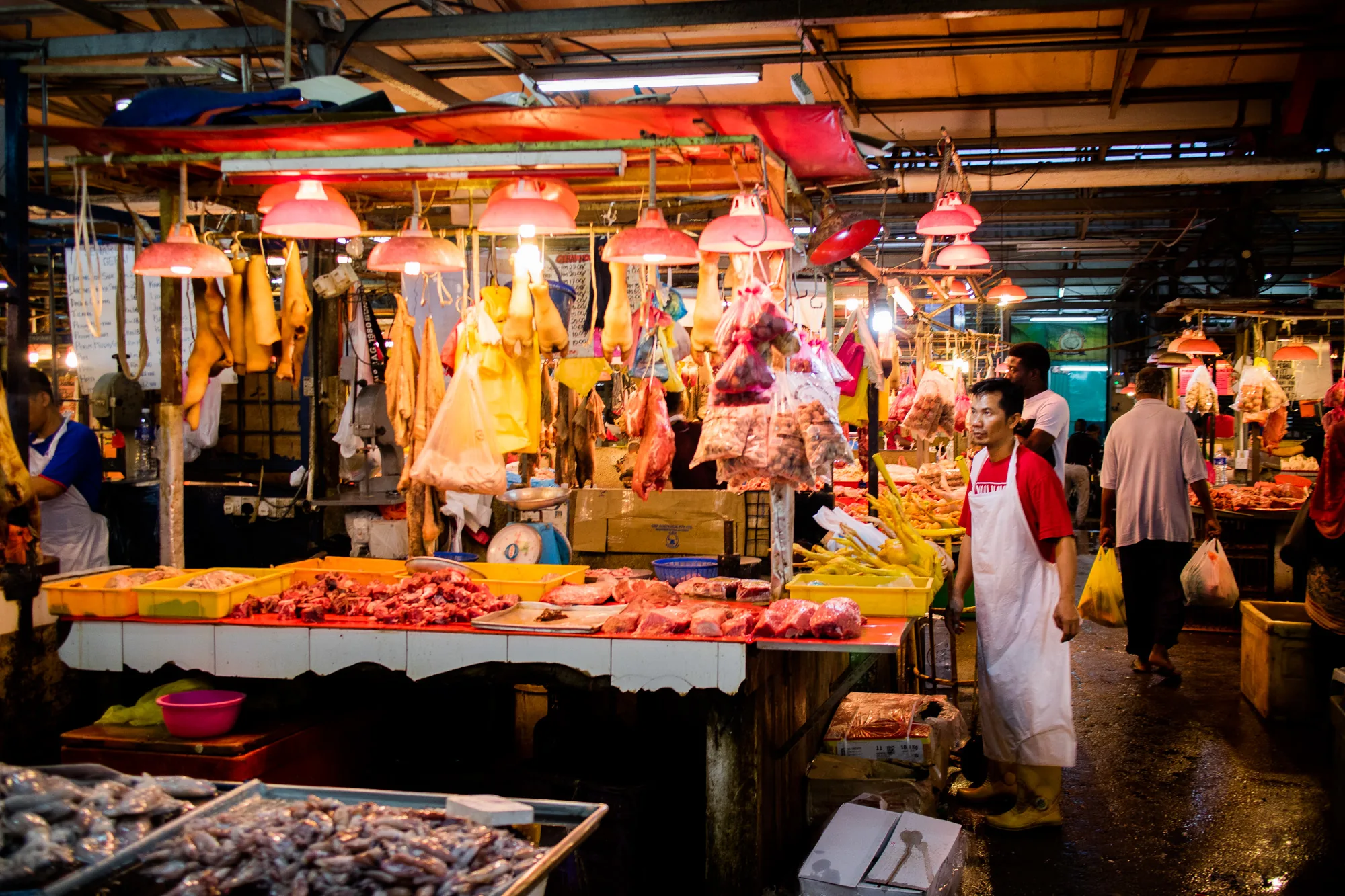 この写真はマレーシアのクアラルンプールの市場の様子を捉えています。薄暗い照明の下、肉や魚介類が所狭しと並べられています。赤色の傘型の照明が吊り下げられ、食品を照らしています。販売員は赤いシャツに白いエプロン、長靴を履いており、市場の活気を感じさせます。床は濡れており、商品の入った袋を提げた買い物客が行き交い、賑わっています。吊るされた肉や鶏肉、新鮮な魚介類など、多様な食材が豊富に揃っていることが伺えます。