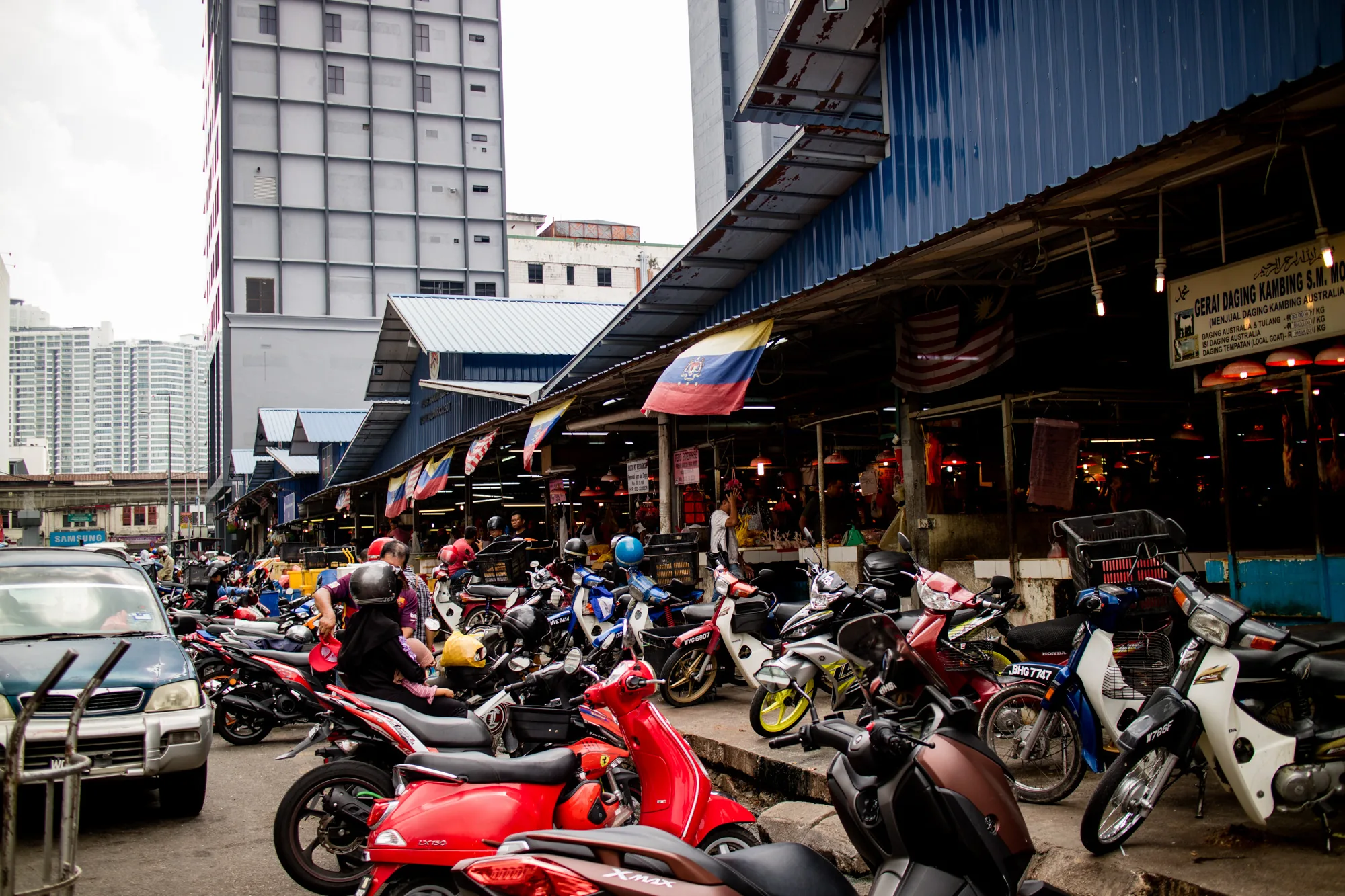 写真はマレーシアのクアラルンプールで撮影されたもので、活気ある市場の様子を捉えています。画面手前には、赤、黒、茶色など様々な色のバイクが多数駐車されており、人々の生活の一部となっていることが伺えます。市場の建物は青い屋根で覆われており、マレーシア国旗や他の旗が飾られています。建物の中は薄暗く、様々な商品が並んでいる様子が見えます。背景には高層ビル群がそびえ立ち、都市の発展を感じさせます。曇り空の下、市場は活気があり、人々の生活感が伝わってくる写真です。