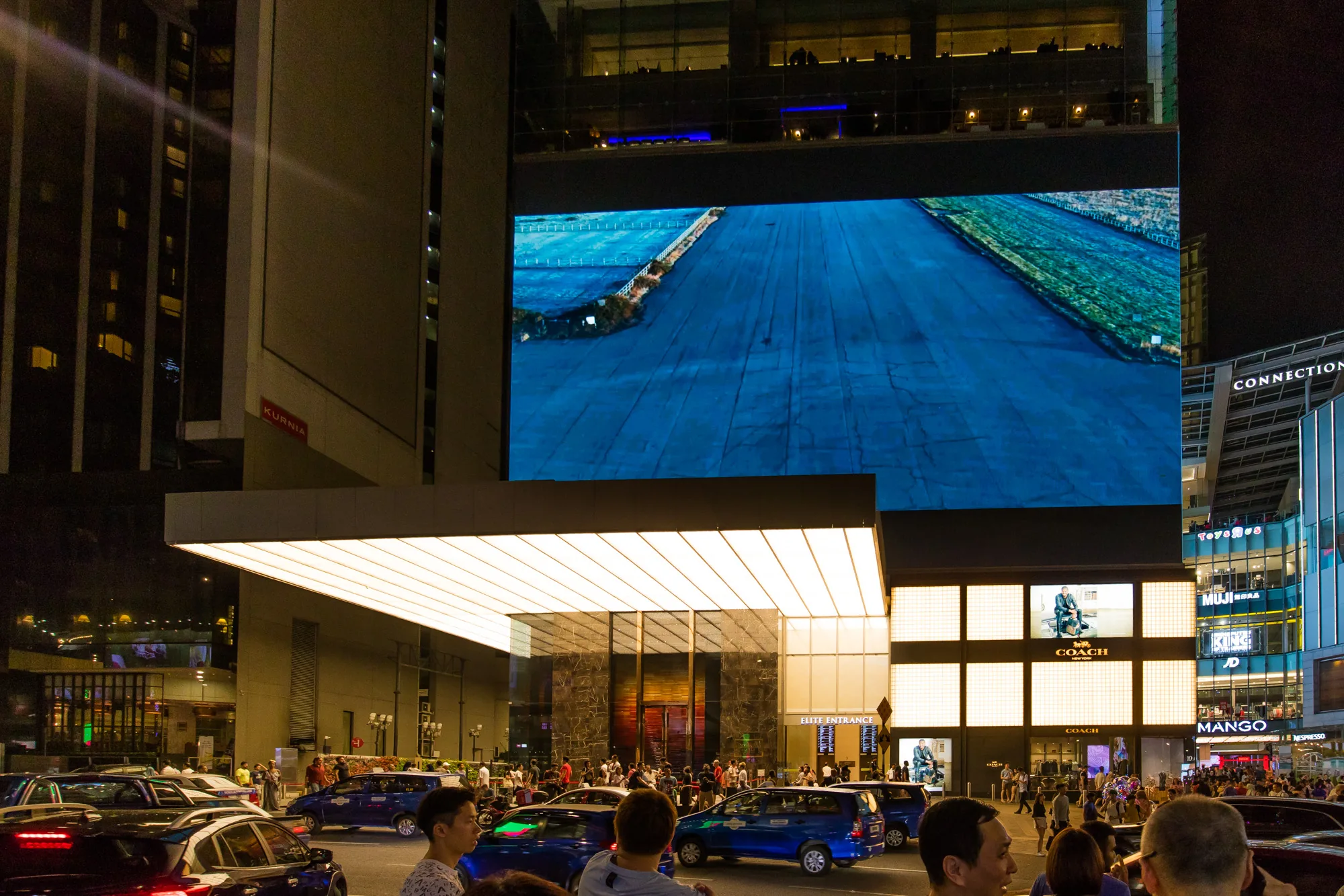 写真はマレーシアのクアラルンプールで撮影された夜景です。 高層ビルが立ち並び、ネオンや照明が輝いています。 特に目を引くのは、巨大な LED スクリーンに映し出された映像です。 道路には車や人が行き交い、賑やかな雰囲気です。 COACHなどのブランドの広告も見られます。 全体的に、現代的な都市の活気が感じられる一枚です。