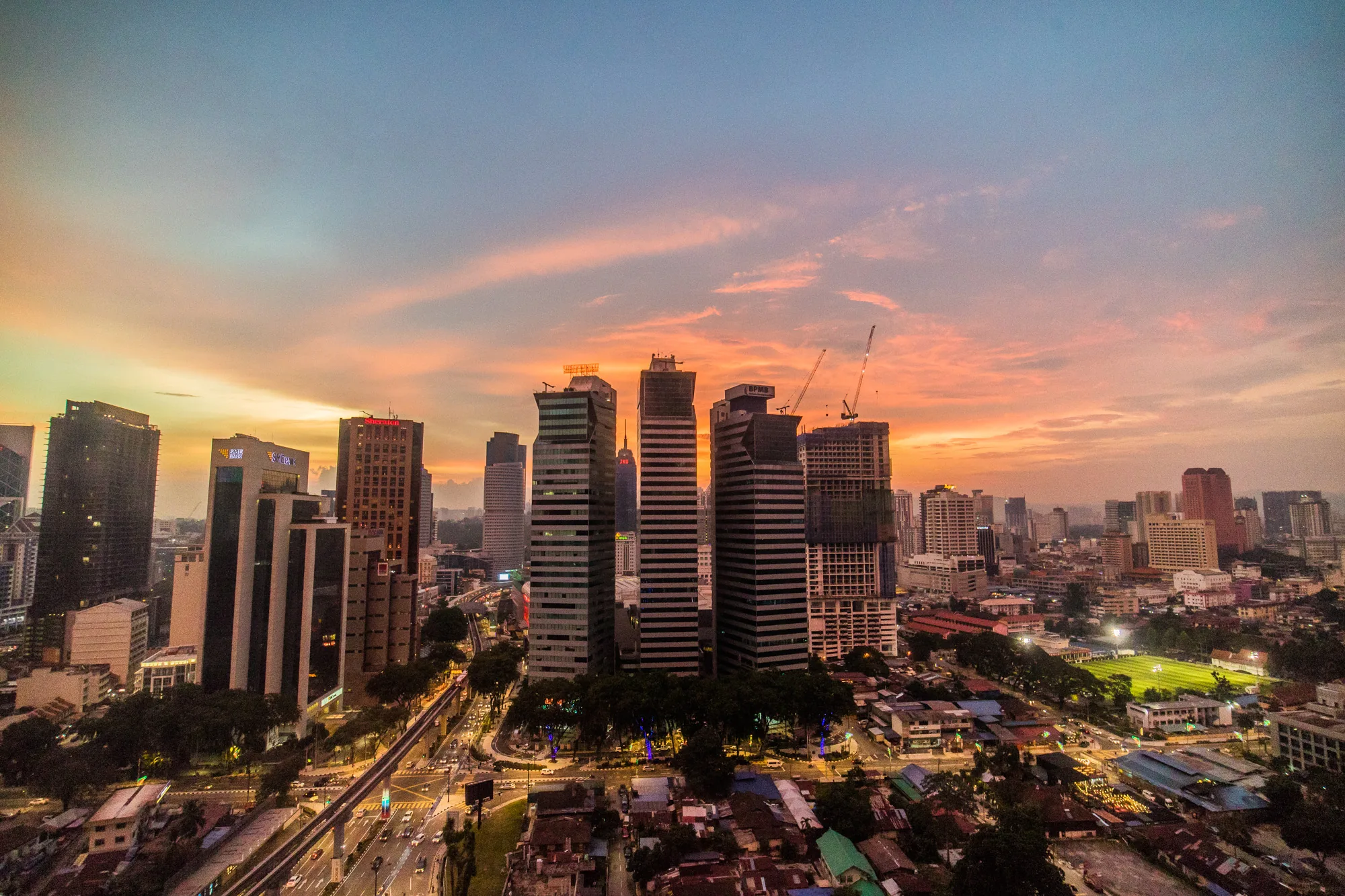 マレーシアのクアラルンプールで撮影された、夕暮れ時の都市景観の写真です。空は青色からオレンジ色へとグラデーションを描き、薄い雲が夕日に染まってピンク色に輝いています。画面中央には高層ビル群がそびえ立ち、そのシルエットが夕焼け空に映えています。いくつかのビルには、建設中のクレーンが見えます。  ビルの下には、街並みが広がっています。道路には車が走り、ヘッドライトの光跡が街の活気を伝えています。緑豊かな木々が点在し、都市の中に自然が共存している様子が伺えます。写真全体が暖色系の光に包まれており、都会の喧騒と夕暮れの穏やかさが調和した美しい風景です。