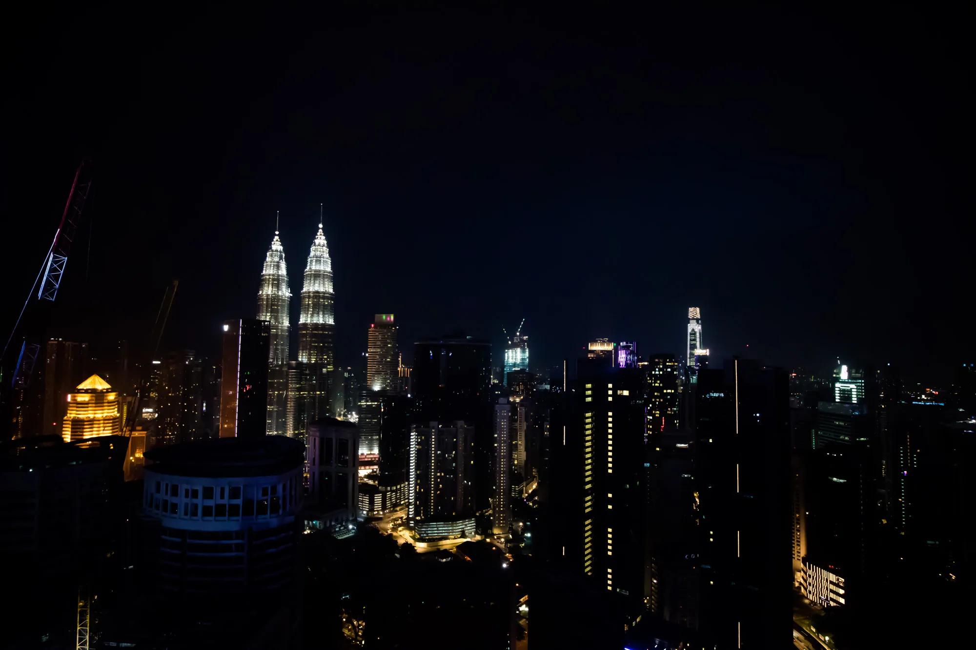 マレーシアのクアラルンプールで撮影された夜景の写真です。特にペトロナスツインタワーが明るく輝き、ひときわ目を引きます。周囲の建物も様々な色や形でライトアップされており、複雑な都市のシルエットを形作っています。建設中のクレーンもいくつか見られ、都市の開発が進んでいる様子が伺えます。全体的に暗い夜空を背景に、人工的な光が鮮やかに映える、幻想的な雰囲気の写真です。