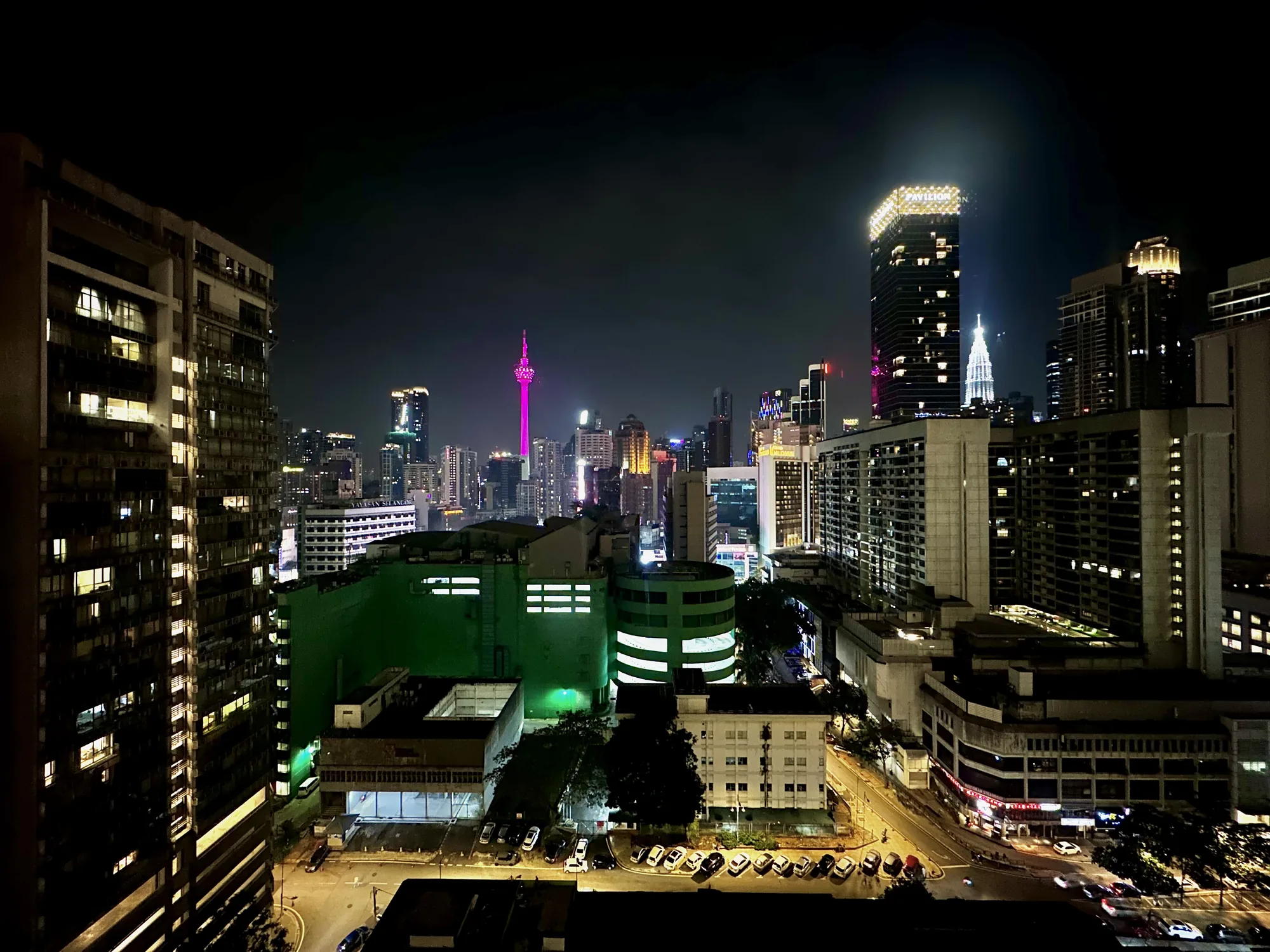 この写真は、マレーシアのクアラルンプールで撮影された夜景です。高い場所から見下ろした都市の風景が広がっており、暗い空の下、無数の高層ビルがライトアップされています。  写真の中央には、鮮やかなピンク色にライトアップされたクアラルンプールタワーが際立っており、夜空にそびえ立っています。その近くには、白く輝くペトロナスツインタワーも確認できます。他の高層ビルも様々な色でライトアップされており、都市の活気と多様性を感じさせます。  手前には、緑色の建物や駐車場、街路灯に照らされた道路が見えます。駐車場には多くの車が停まっており、道路には車のライトが点々と続いています。写真全体からは、夜のクアラルンプールの賑やかさと美しい景観が伝わってきます。