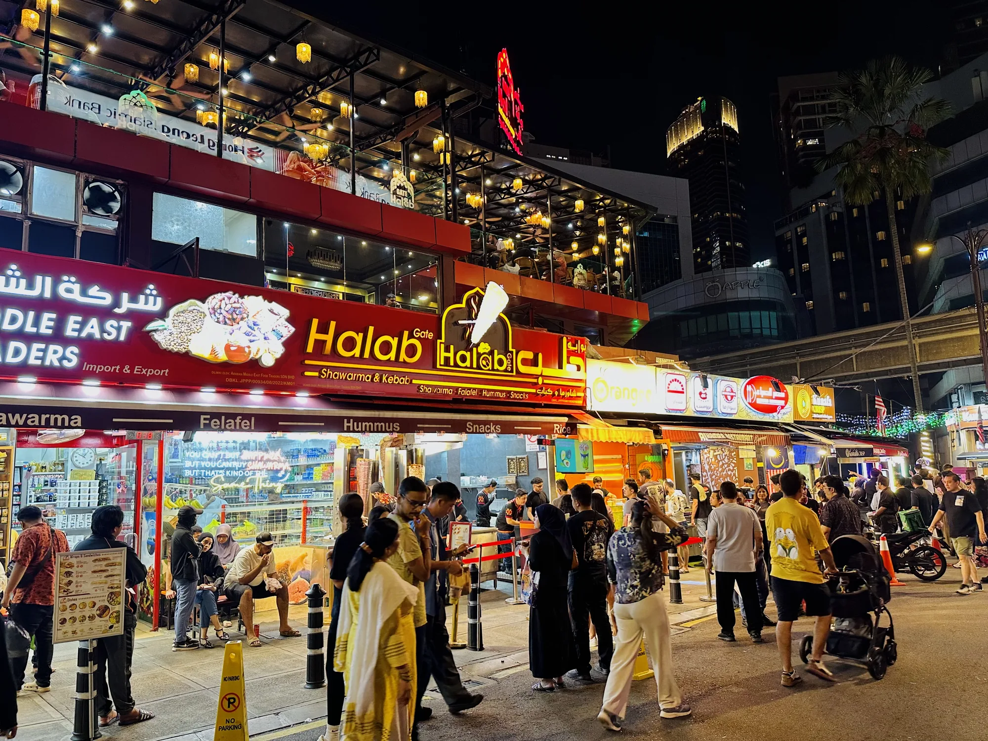 この写真は、マレーシアのクアラルンプールで撮影された夜の街の様子を捉えています。通りには多くの人々が行き交い、活気に満ち溢れています。写真の中心には、アラビア文字が書かれた看板を掲げた飲食店が立ち並んでいます。看板には「Halab」という文字が見え、シャワルマやケバブ、ファラフェルなどを提供していることがわかります。他にも、「Middle East Traders」と書かれた看板や、メニューが描かれた立て看板などがあり、様々な飲食店が軒を連ねている様子が伺えます。  通りの人々は、様々な服装をしており、家族連れや友人同士など、多様なグループが見られます。中には、ベビーカーを押す人や、テイクアウトの料理を受け取る人もいます。通りの奥には、高層ビルがそびえ立ち、ネオンサインや街灯が夜空を照らしています。建物の壁面には「APPLE」のロゴも見えます。全体的に、賑やかで異国情緒漂う雰囲気が感じられる写真です。