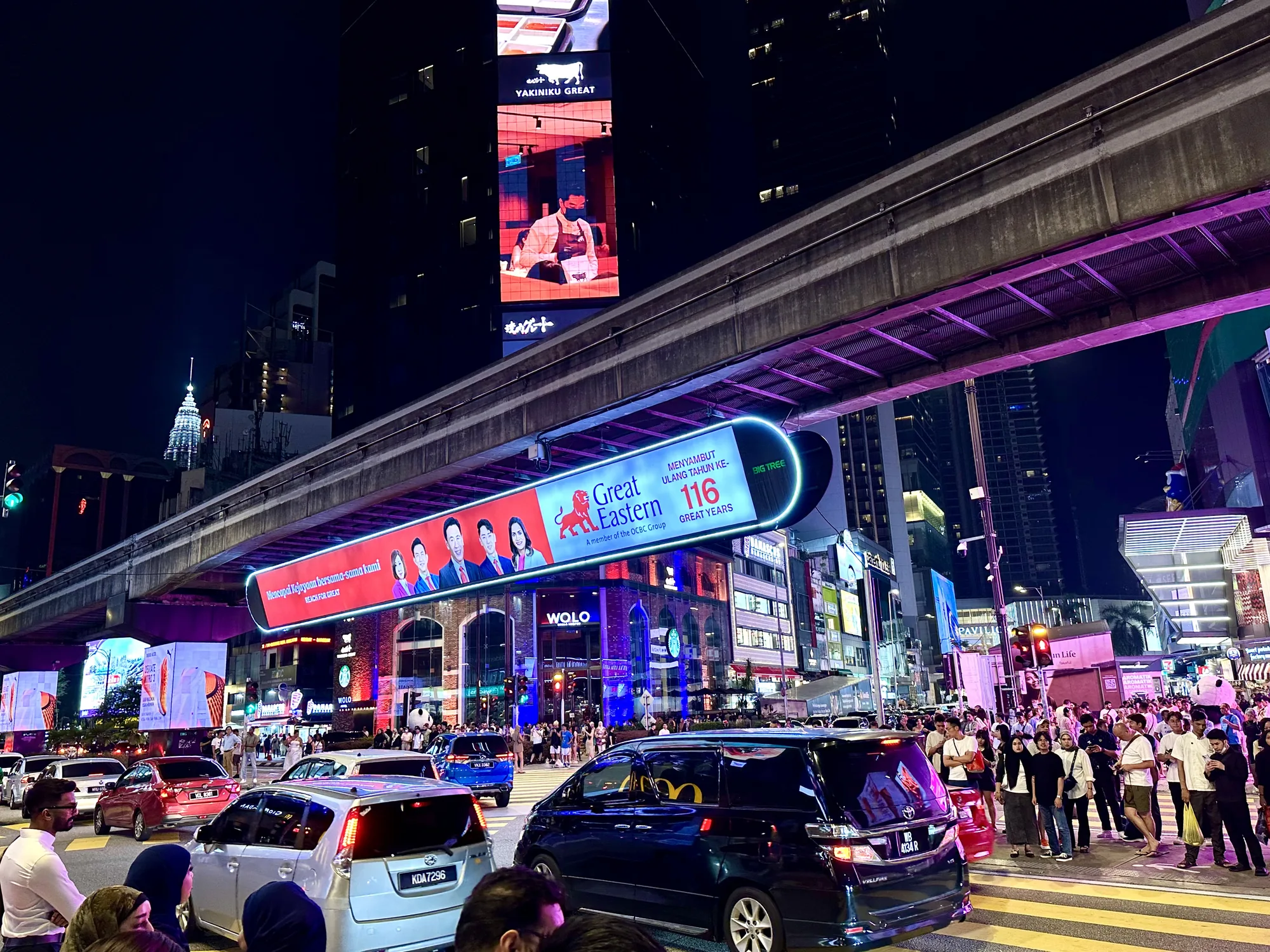 写真はマレーシアのクアラルンプールで撮影された夜の街並みを捉えています。道路は車で混雑しており、歩道には多くの人が行き交っています。高架鉄道が頭上を走り、その下には巨大な広告が表示されています。"Great Eastern"という企業の広告で、創業116周年を祝っているようです。背の高いビルには"YAKINIKU GREAT"という焼肉店の広告が出ています。街全体が明るく、活気があり、賑やかな雰囲気です。