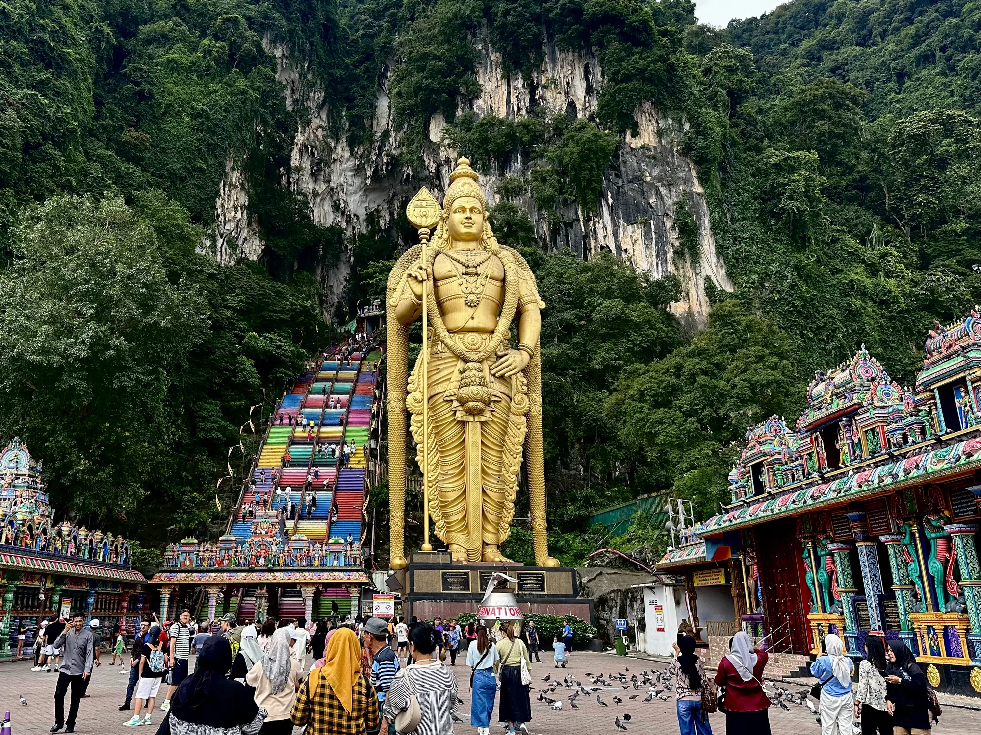 マレーシアのクアラルンプールにあるバトゥ洞窟を撮影した写真です。画面中央には、巨大な黄金のムルガン像がそびえ立っています。像の背後には、洞窟へと続くカラフルな階段が伸びており、多くの観光客が階段を上り下りしています。階段の左右には緑豊かな木々が生い茂り、自然の美しさを際立たせています。画面下部には、多くの人々が集まっており、観光地の賑わいを伝えています。また、地面には鳩が群れており、のどかな雰囲気を醸し出しています。右側には、色鮮やかな装飾が施された寺院の建物が見えます。全体的に、バトゥ洞窟の壮大さと活気が感じられる写真です。