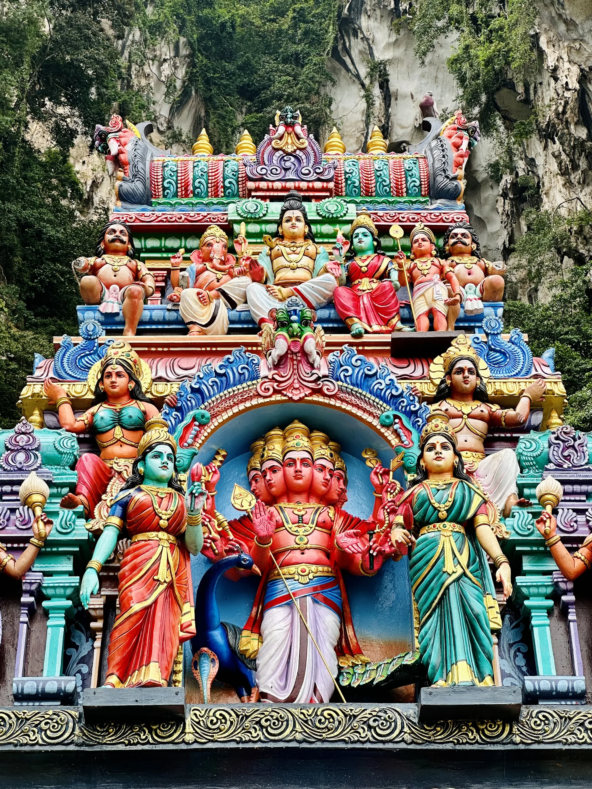 写真はマレーシアのクアラルンプールで撮影された寺院の彫刻のクローズアップです。画面全体に、鮮やかな色彩で彩られたヒンドゥー教の神々の像が積み重なるように配置されています。一番下の段には、複数の顔と腕を持つ神の像があり、その両脇にはそれぞれ異なる色の衣装をまとった女神像が立っています。像の隣には孔雀が彫刻されています。その上の段には、さらに多くの女神像が並んでいます。最上段には、男性神と女性神、ガネーシャ神など、多様な神々が座っている様子が表現されています。彫刻全体は複雑な模様で装飾されており、背景には岩山と緑が生い茂っています。写真全体に鮮やかな色彩と細やかな装飾が施されており、宗教的な雰囲気と異国情緒が感じられます。
