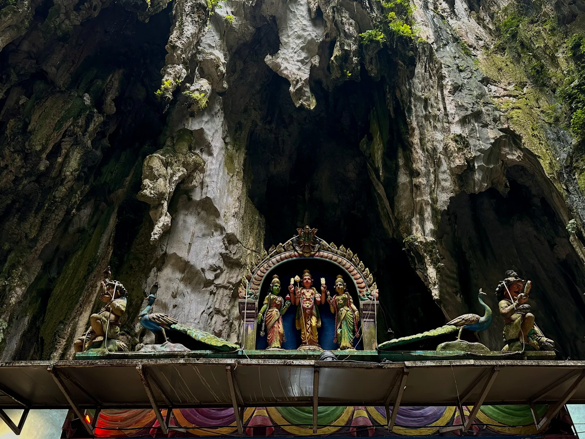写真はマレーシアのクアラルンプールにあるバツー洞窟で撮影されたものです。石灰岩の洞窟の入り口に、鮮やかな色の神像が飾られています。洞窟の岩肌はゴツゴツとしており、ところどころ緑の植物が生えています。神像は精巧に作られており、中央には鮮やかな衣装をまとった3体の神様が並んでいます。神像の左右には、クジャクや人物の像が配置されており、さらにその上には小さな鳥が止まっています。全体的に、洞窟の荘厳さと神聖な雰囲気が伝わってくる写真です。洞窟内部の暗さと、神像の鮮やかな色彩のコントラストが印象的です。