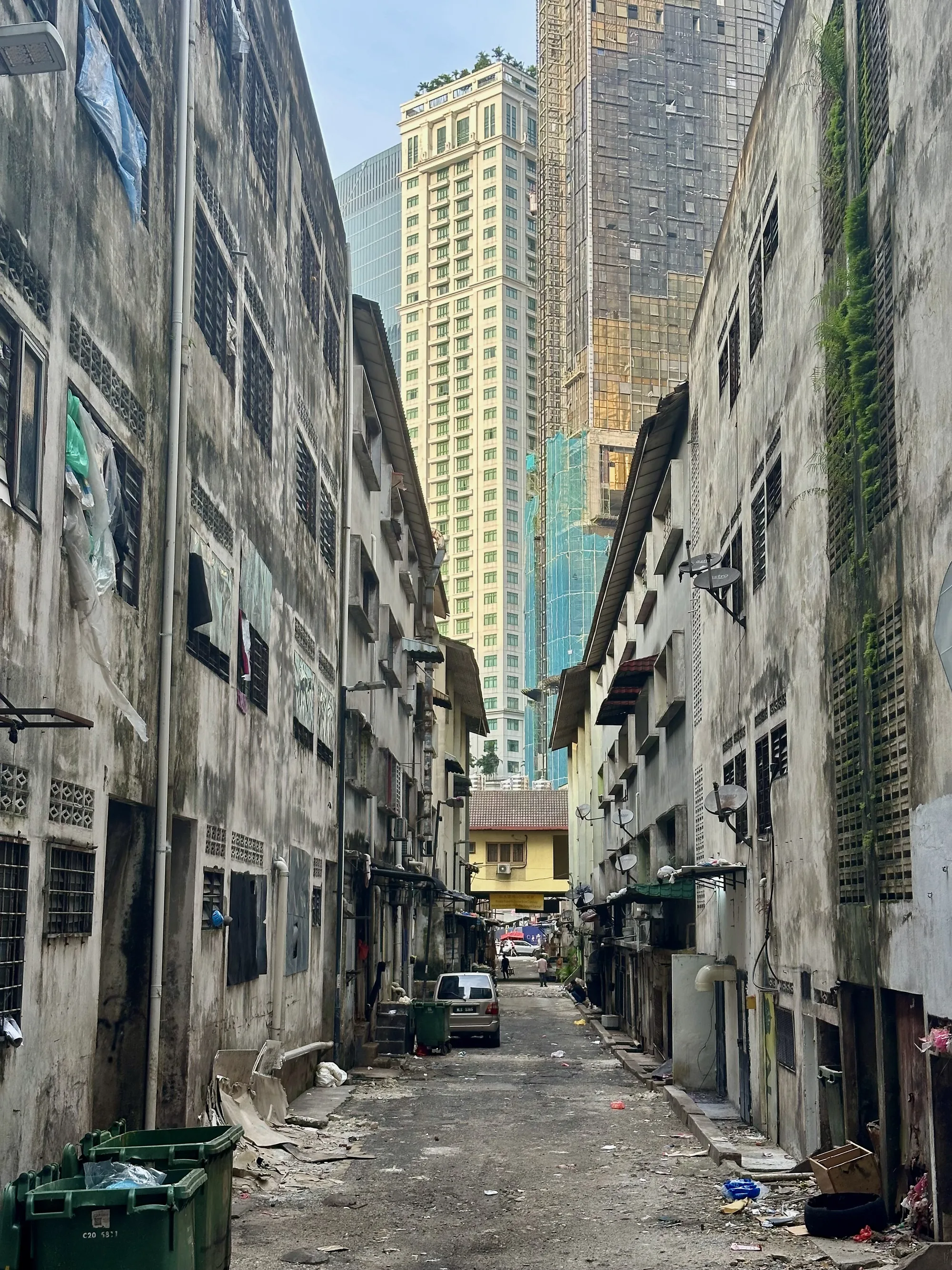 この写真は、マレーシアのクアラルンプールで撮影されたものと思われます。写真には、古びた建物が立ち並ぶ路地と、その後ろにそびえ立つ近代的な高層ビル群が写っており、強いコントラストが印象的です。  路地は狭く、道の表面は荒れており、ゴミが散乱している箇所も見られます。両側の建物は古く、壁は汚れや苔で覆われており、歴史を感じさせます。窓には格子がはめられ、一部には洗濯物が干されていたり、ビニールシートで覆われているものもあります。建物の外壁にはエアコンの室外機や衛星アンテナが取り付けられており、生活感があります。  しかし、路地の奥には、光り輝く高層ビル群がそびえ立っており、近代的な都市の発展を象徴しています。特に、白くスタイリッシュな高層ビルは、屋上に緑が植えられており、都会のオアシスのような雰囲気を醸し出しています。建設中の高層ビルもあり、都市の成長のスピードを感じさせます。  全体的に、この写真は貧困と富、過去と未来が混在するクアラルンプールの複雑な側面を捉えているように感じられます。路地の生活感あふれる様子と、高層ビルの洗練されたデザインが対照的で、見る人に様々な感情を抱かせる一枚です。