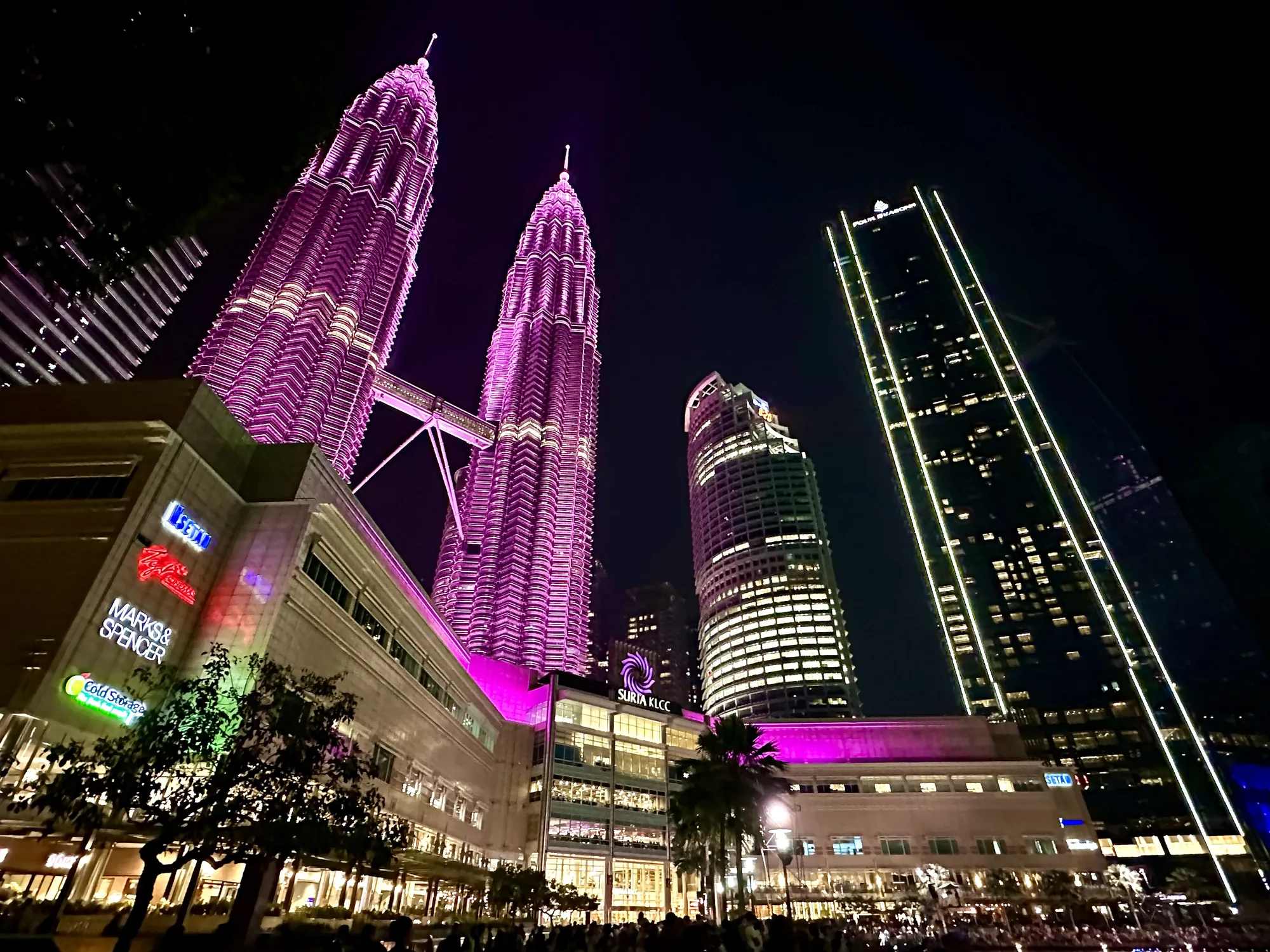 マレーシアのクアラルンプールで撮影された夜景の写真です。画面全体に渡り複数の高層ビルがそびえ立ち、都会の夜の賑わいを表現しています。  ひときわ目を引くのは、紫色のライトアップが施されたペトロナスツインタワーです。2つのタワーは連結されており、その独特な形状と鮮やかな色彩は、クアラルンプールを象徴する景観として際立っています。  ペトロナスツインタワーの手前には、ショッピングモールの「SURIA KLCC」が見えます。モール自体もライトアップされており、賑わいを演出しています。また、「MARKS & SPENCER」や「COLD STORAGE」といった店舗の看板も確認できます。  その他、垂直方向にライトが走る高層ビルや、曲線的なデザインが特徴的な高層ビルも写っており、クアラルンプールの多様な建築様式を垣間見ることができます。  画面下部には、街路樹と、その下を歩く人々のシルエットが写っており、夜の街の活気が感じられます。全体的に、照明が強く、鮮やかな色彩が印象的な写真です。