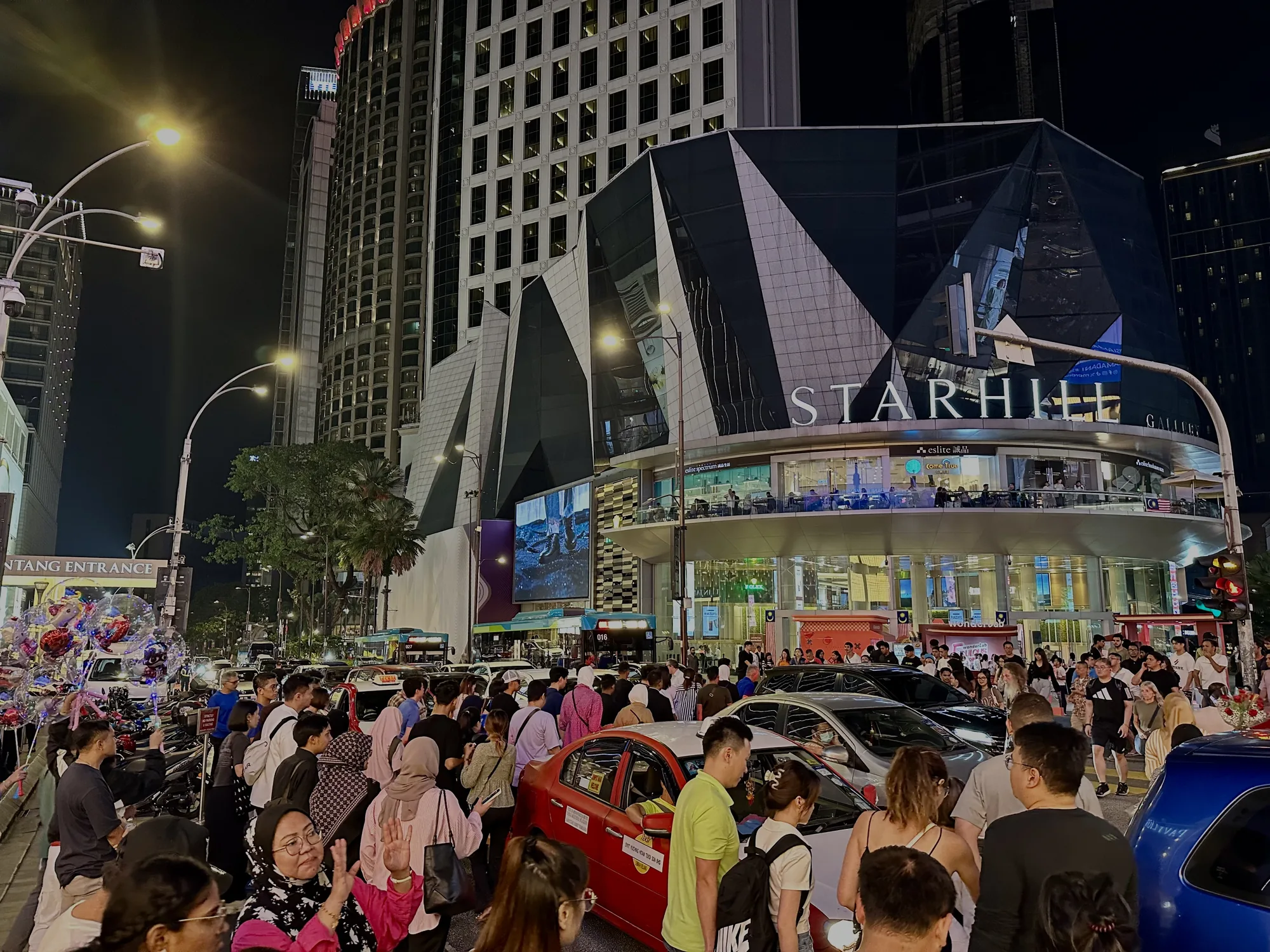 写真はマレーシアのクアラルンプールで撮影された夜景です。スターヒルギャラリー（Starhill Gallery）が大きく写っており、その前には多くの人々がいます。通りには車やバスが走っており、交通量が多いことが伺えます。建物の照明が明るく、夜の街の賑わいを表しています。スターヒルギャラリーの看板がはっきりと確認でき、そのモダンな建築様式が印象的です。また、街灯や建物の明かりが夜空を照らし、活気ある雰囲気を醸し出しています。人々は様々な服装をしており、観光客や地元の人々が混ざり合っている様子が分かります。歩道には風船を持った人がおり、お祭りかイベントがあるのかもしれません。全体的に、クアラルンプールの都市の活気と魅力を伝える写真です。