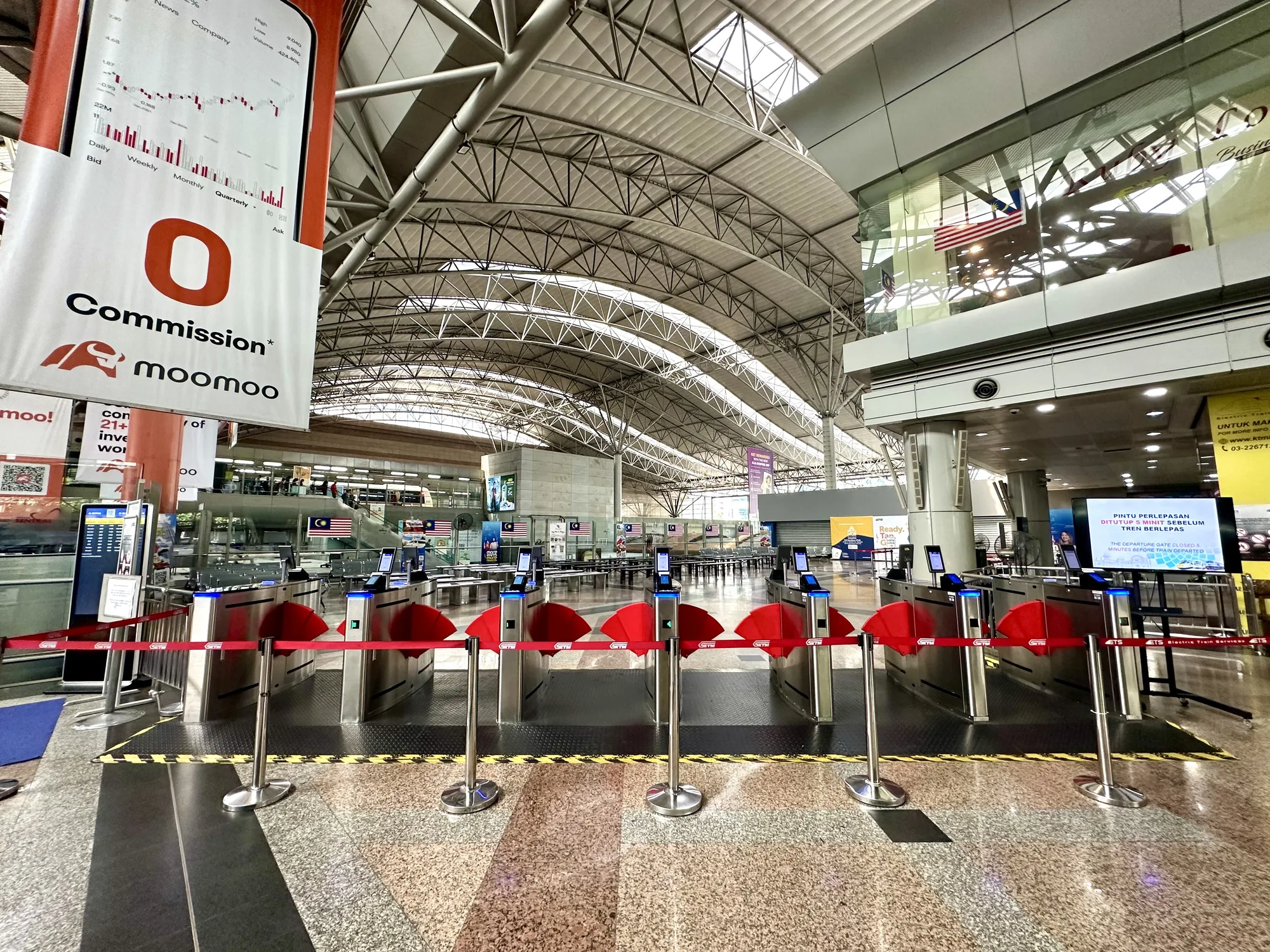 写真はマレーシアのクアラルンプールにある駅の構内を撮影したものです。画面手前には、銀色の自動改札機が複数並んでおり、赤い仕切りが張られています。改札機の上には、青いライトが点灯しています。床はグレーを基調とした模様が施されており、黄色と黒の注意喚起のテープが貼られています。天井は高く、複雑な金属製の構造がむき出しになっています。奥には、マレーシア国旗や広告などが見えます。画面左側には、「moomoo」という金融関連の広告バナーが掲げられています。全体的に、明るく清潔感のある雰囲気で、近代的な駅の様子が伝わってきます。