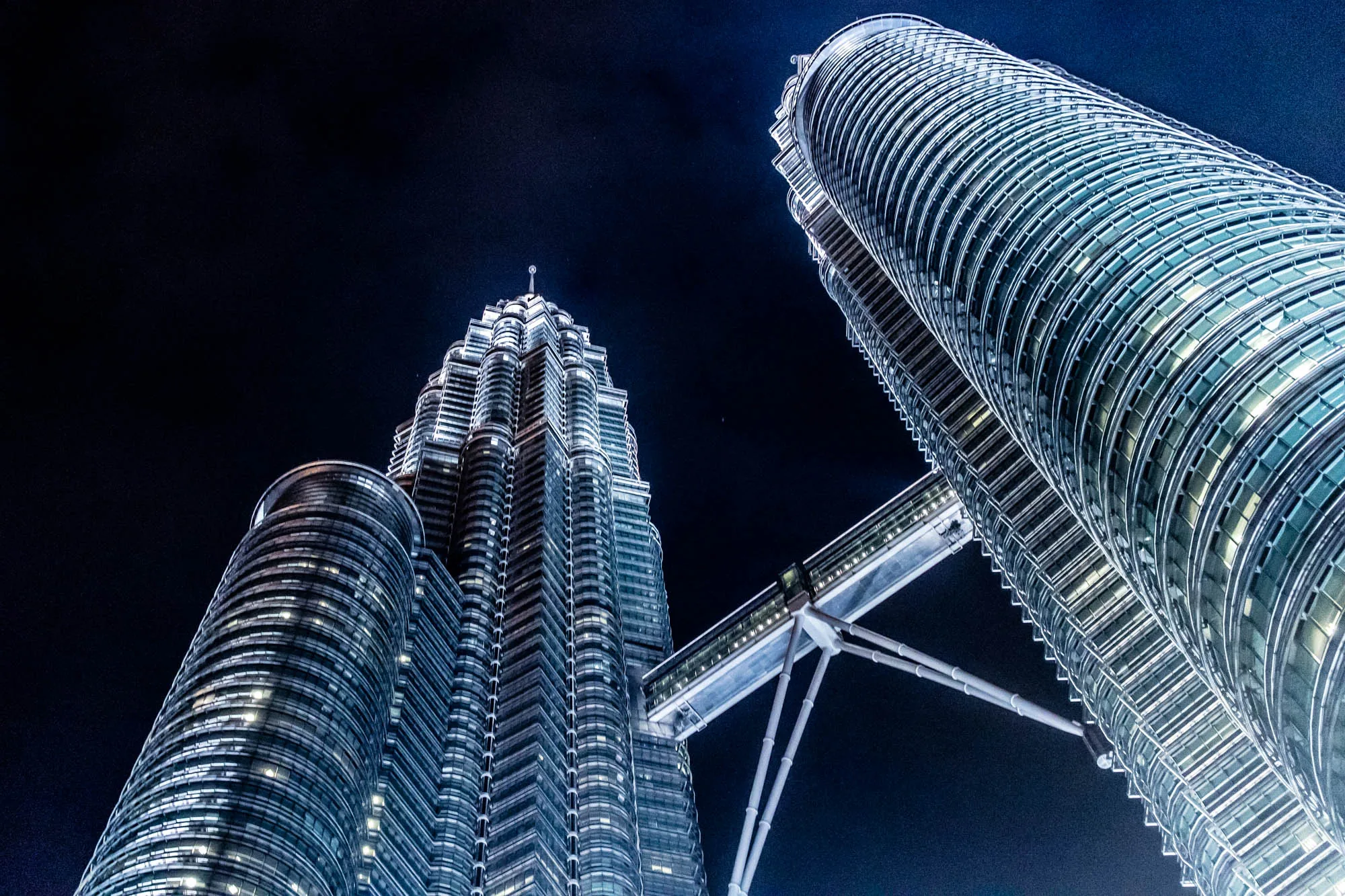 マレーシアのクアラルンプールで撮影された、夜のペトロナスツインタワーの写真です。見上げるようなローアングルから捉えられており、夜空を背景に、2つのタワーがそびえ立っています。タワーは明るくライトアップされており、その幾何学的な構造が際立っています。2つのタワーを結ぶスカイブリッジもはっきりと見え、近代的な建築美を感じさせます。全体的に青みがかった光が印象的で、クールで都会的な雰囲気を醸し出しています。夜空には、小さな星がいくつか見えます。