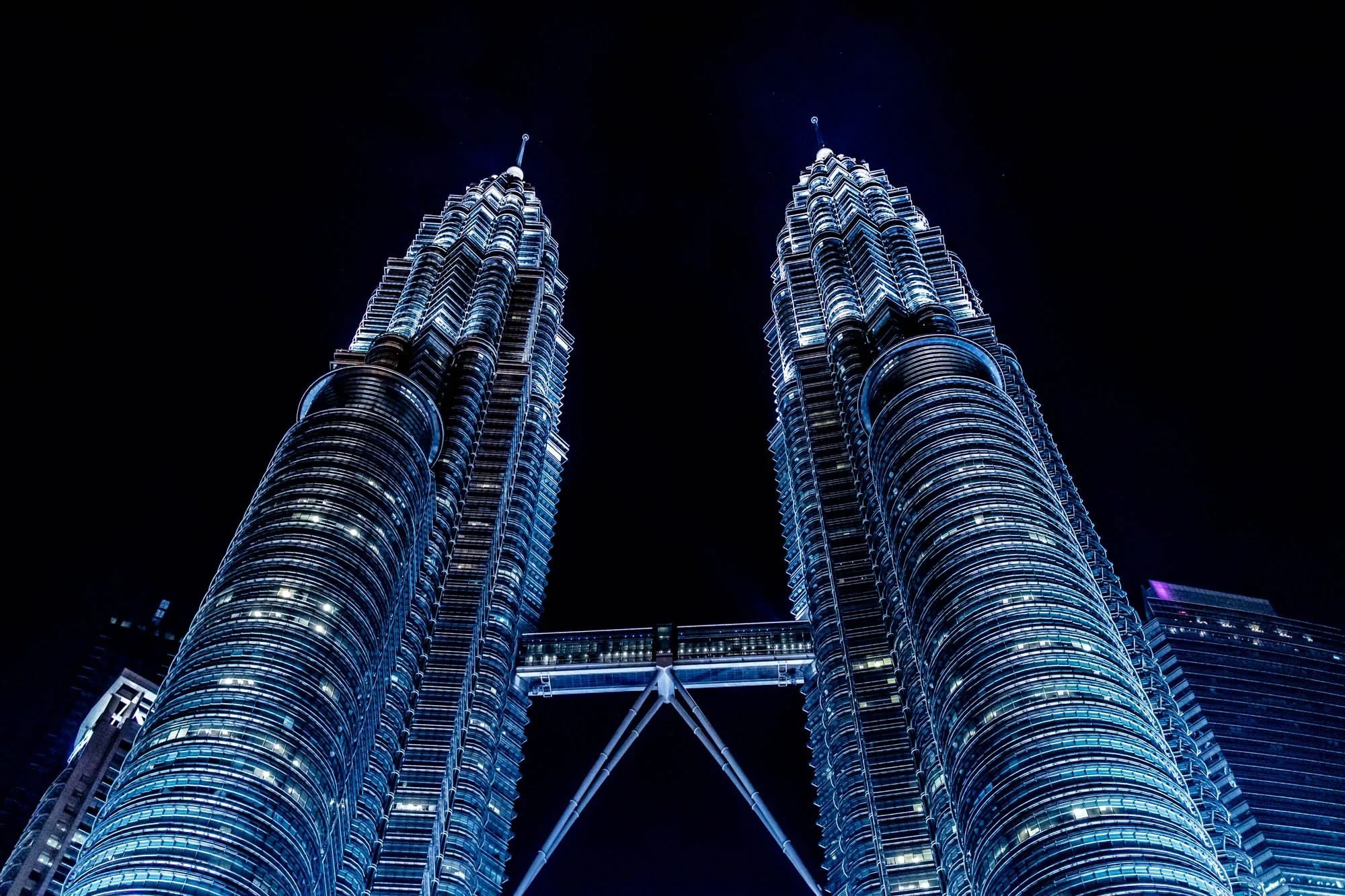 クアラルンプールの夜景を捉えた写真です。画面いっぱいにそびえ立つのは、マレーシアの象徴とも言えるペトロナスツインタワーです。夜空を背景に、二つのタワーが並び立っています。タワー全体が青白い光でライトアップされており、無数の窓から漏れる光が、まるで夜空に輝く星のように見えます。二つのタワーを結ぶスカイブリッジもライトアップされ、独特の存在感を放っています。下から見上げるようなアングルで撮影されており、タワーの巨大さ、壮大さが強調されています。画面全体は暗い色調で統一され、夜の静寂さと、タワーの神秘的な雰囲気を醸し出しています。右側には他の高層ビルの一部が写っており、タワーの存在感をより際立たせています。