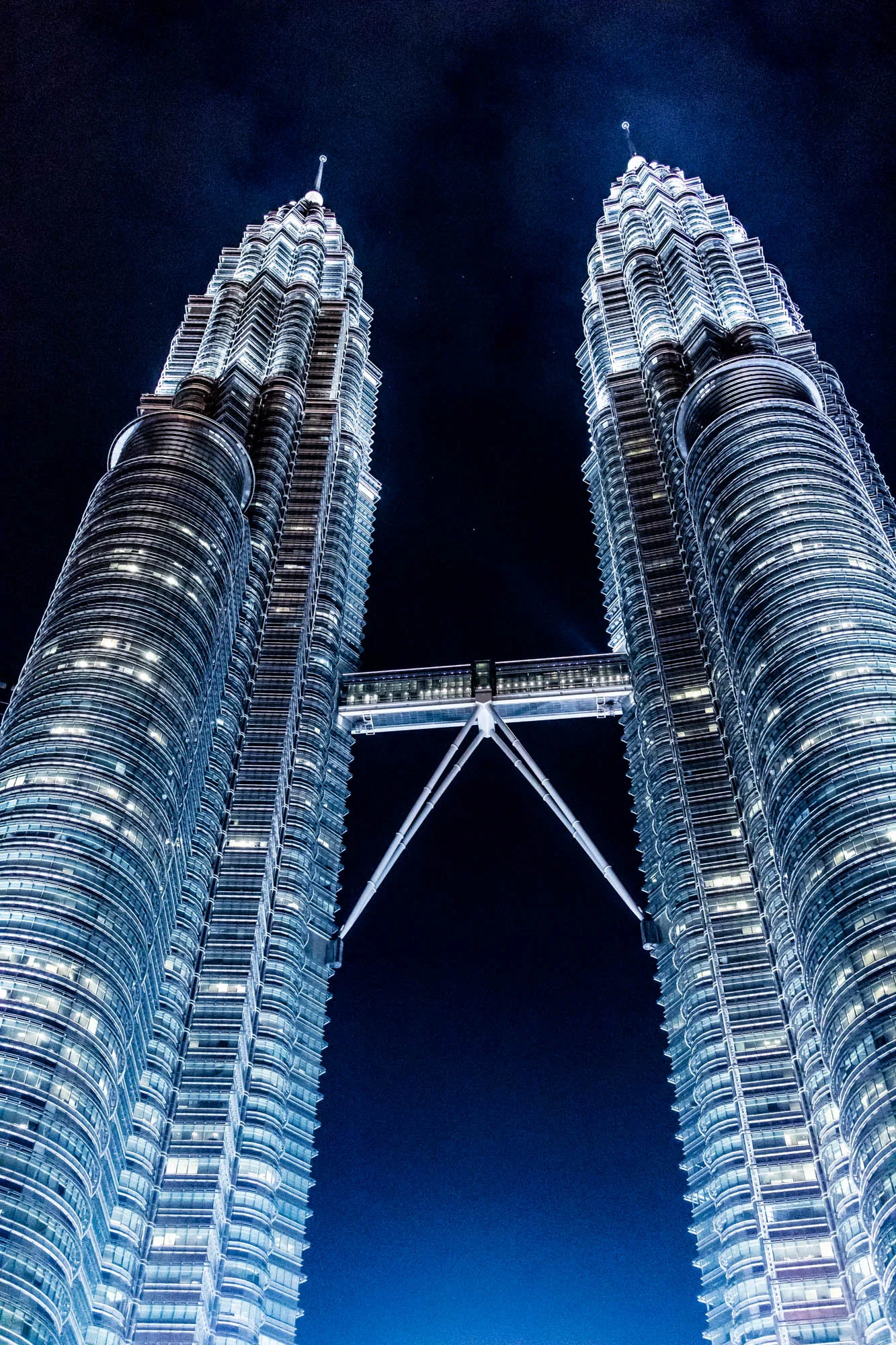マレーシアのクアラルンプールにそびえ立つペトロナスツインタワーを、真下から見上げるように撮影した夜景写真です。暗い夜空を背景に、ライトアップされたツインタワーが圧倒的な存在感を放っています。2つのタワーは、高い位置にあるスカイブリッジで結ばれており、その幾何学的な構造が夜空に浮かび上がります。タワーの表面は無数の窓で覆われ、内部の光が点在し、夜景に独特の輝きを添えています。見上げる構図によって、タワーの高さとスケールの大きさが強調され、見る者に畏敬の念を抱かせます。全体的に青みがかった色調で、クールで都会的な印象を与えます。