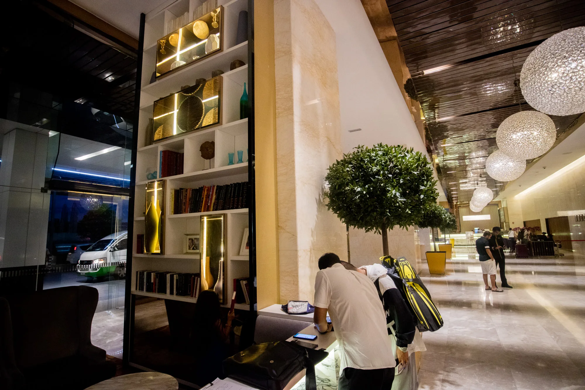 この写真は、マレーシアのクアラルンプールにあるホテルのロビーで撮影されたものです。 ロビーは豪華で、光沢のある大理石の床が印象的です。天井には、球体のシャンデリアが複数吊り下げられており、きらびやかな光を放っています。 画面左側には、装飾品や本が飾られた棚が設置されています。棚の横には大きなガラスがあり、外の様子が少し見えます。 中央には、丸く刈り込まれた緑の木がいくつか配置されており、ロビーに自然な雰囲気を加えています。 手前の受付カウンターには、数人の人が集まっています。一人は携帯電話を見ているようです。 奥の方には、さらに人がいて、ロビー全体が活気に満ちている様子が伺えます。 全体的に、洗練された雰囲気と、活気ある賑わいが感じられる写真です。