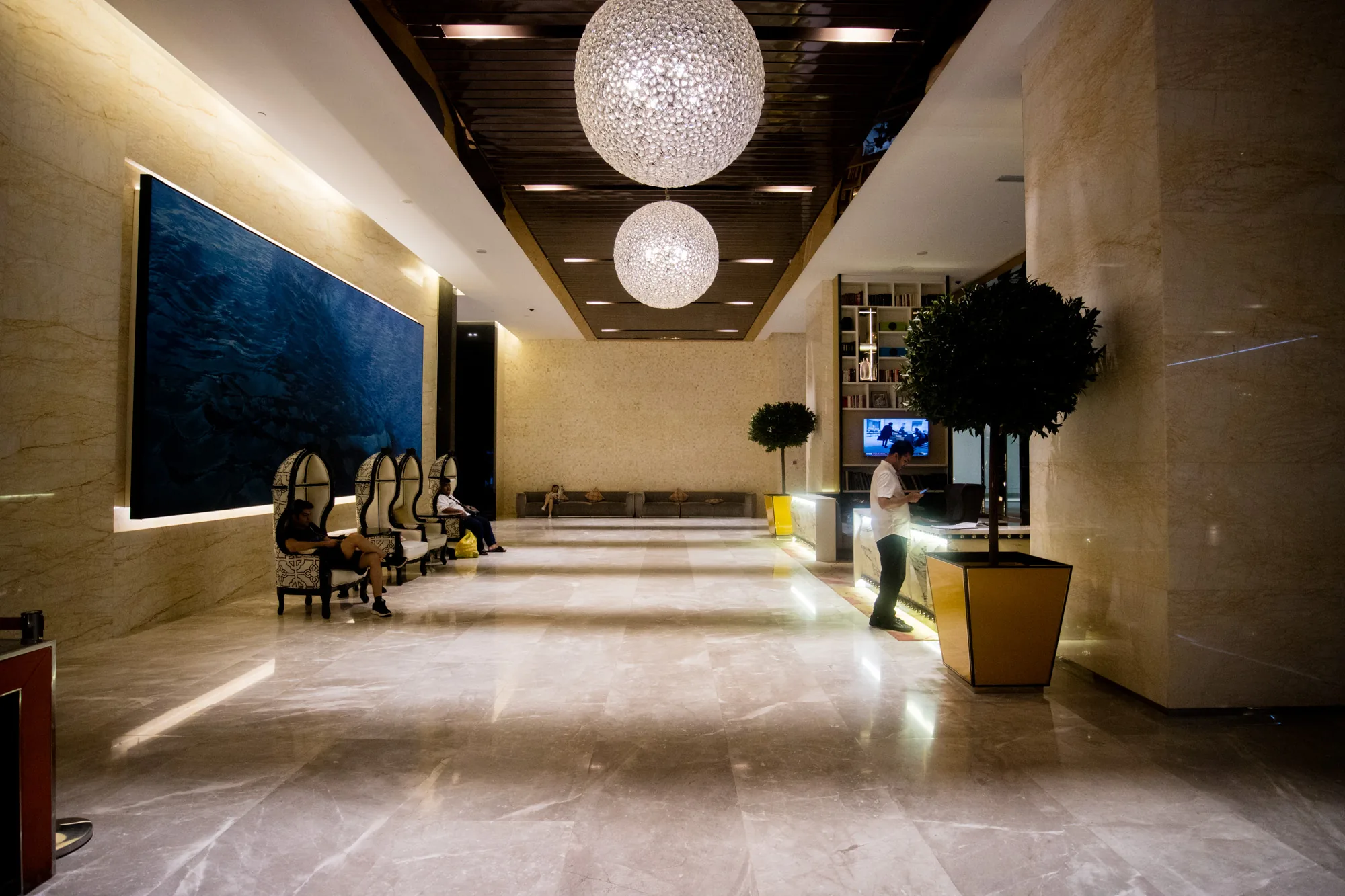 この写真は、マレーシアのクアラルンプールで撮影されたと思われる、高級ホテルのロビーの様子を捉えています。広々とした空間は、光沢のある大理石の床で覆われ、洗練された雰囲気を醸し出しています。  天井からは、キラキラと輝く大きな球体のシャンデリアが二つ吊り下げられており、ロビー全体を明るく照らしています。左側の壁には、鮮やかな青色の巨大なアート作品が飾られており、まるで深海を切り取ったかのような印象を与えます。アート作品の前には、特徴的なデザインの椅子が並んでおり、人々がくつろいで座っています。  ロビーの奥には、ソファが配置された待合スペースがあり、さらに奥には、棚に本が並んだエリアが見えます。また、観葉植物が置かれた受付カウンターの前に、男性が立ってスマートフォンを操作しています。  全体的に、高級感と落ち着きのある空間で、旅行者やビジネスマンがリラックスしたり、待ち合わせをしたりするのに適した場所のように見えます。洗練されたデザインと豪華な装飾が、特別な体験を演出している印象です。